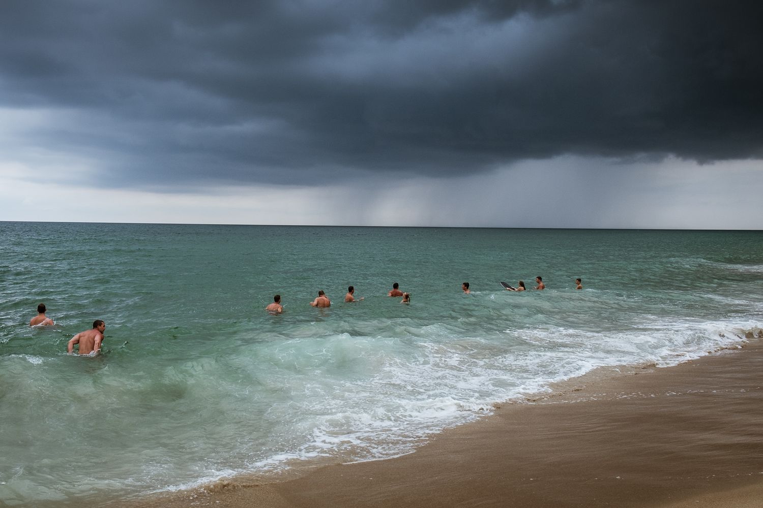 Dramatic stormy clouds gather over a beach with crashing waves and swimmers in the distance.
