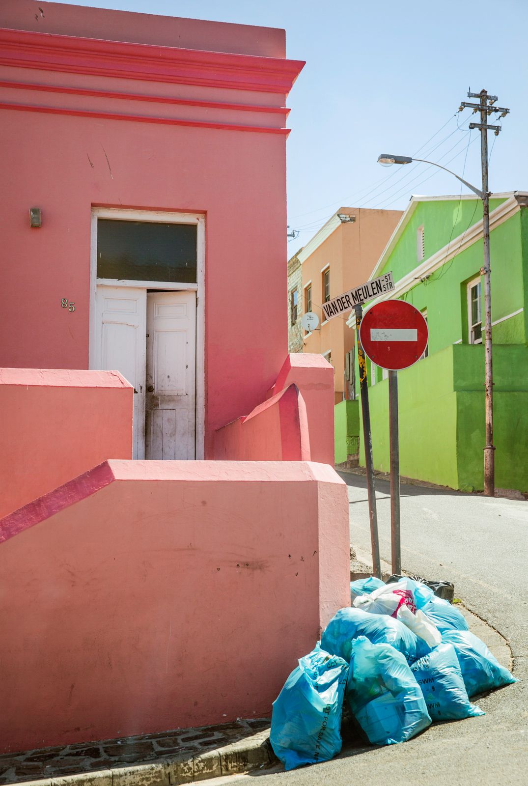 Pink and green buildings with blue garbage bags lined up against wall on urban street.