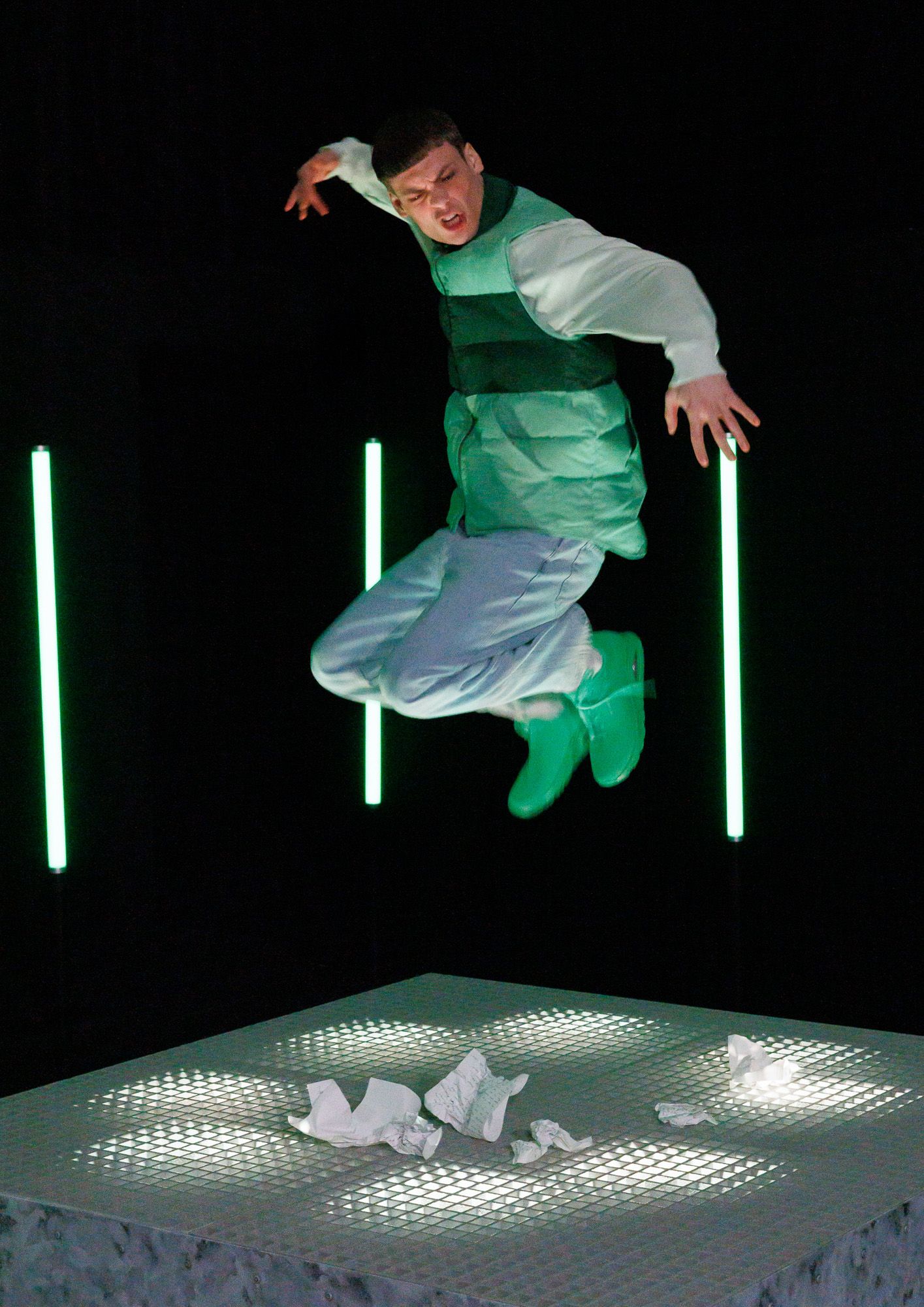 Person in green and white outfit jumping over crumpled paper with neon lights in dark studio setting.