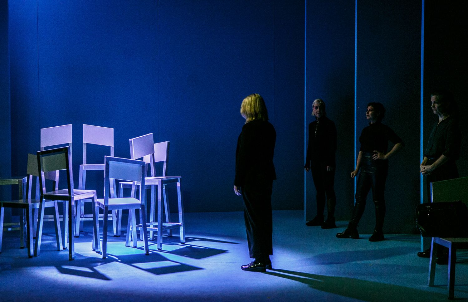 Minimalist theater stage with scattered white chairs under dramatic blue lighting.