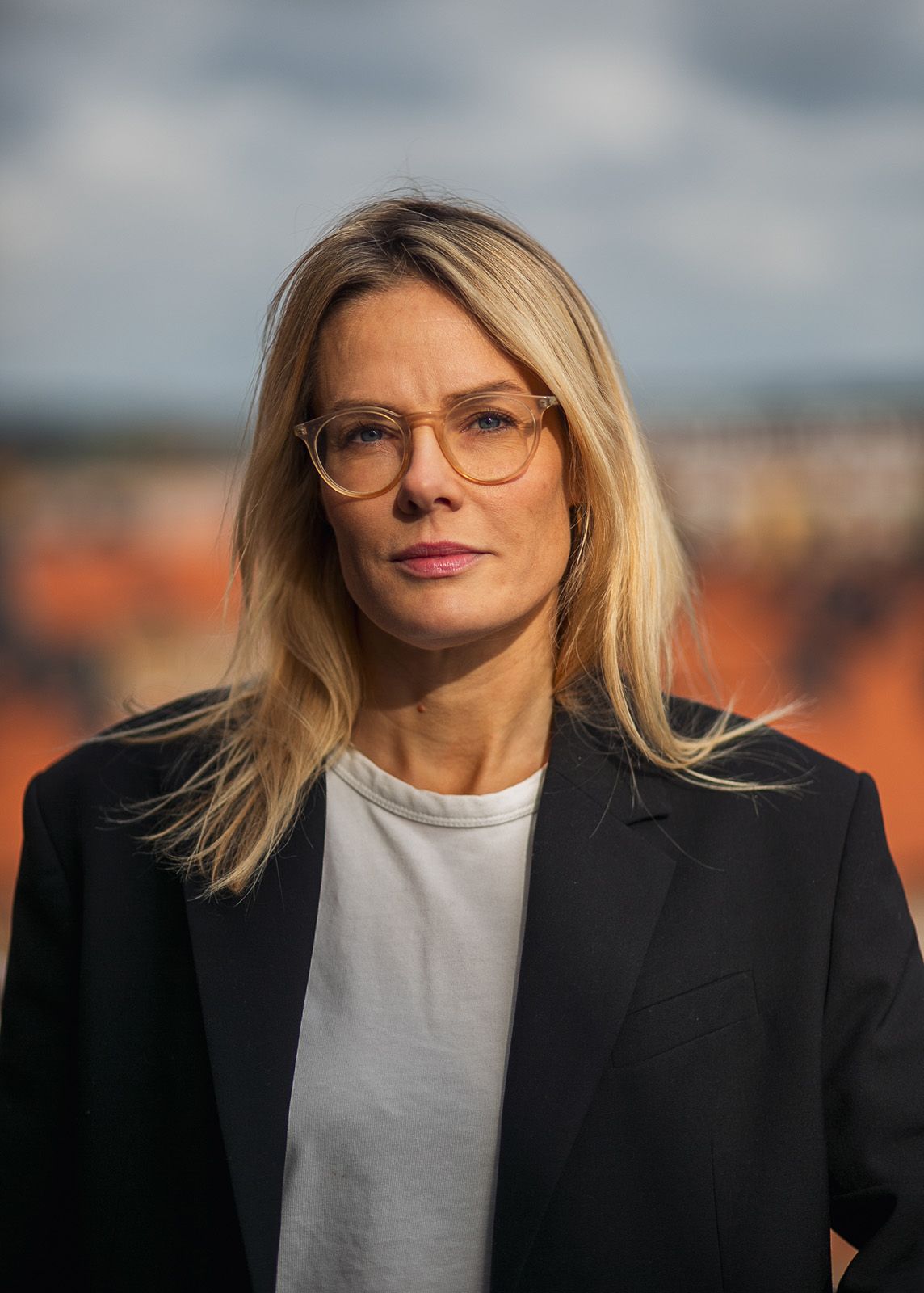 Professionellt porträtt av person i svart kavaj och glasögon mot suddig stadsbildsbakgrund