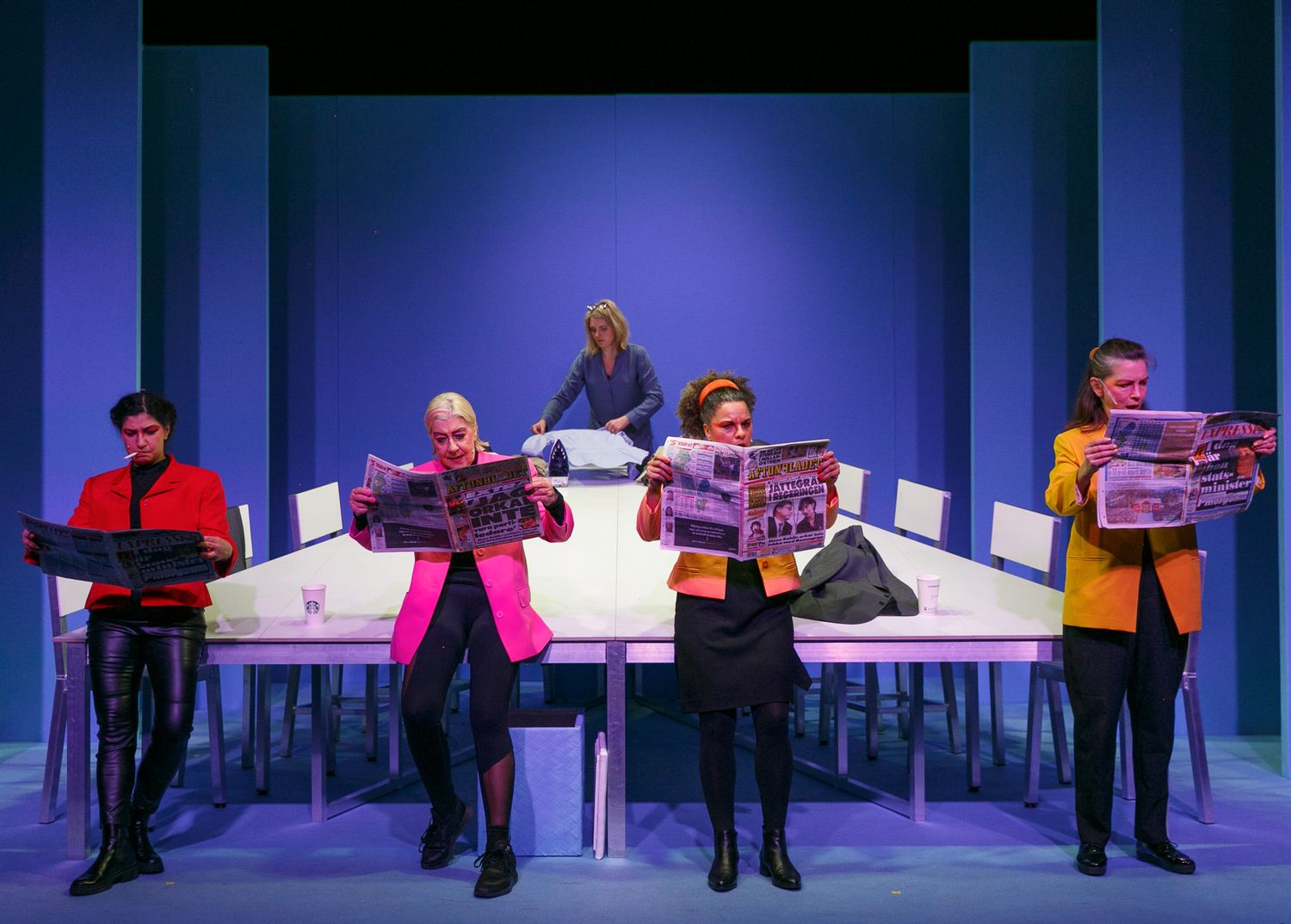 Performers seated on a long bench against a blue backdrop in colorful attire.