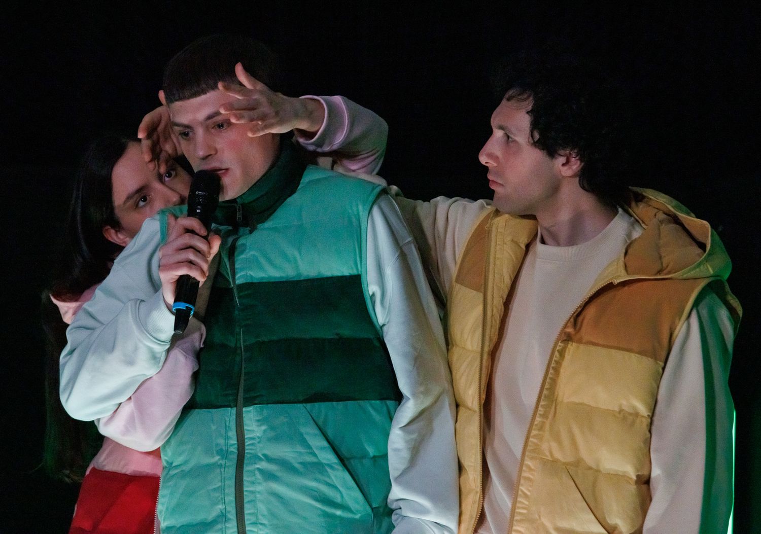 Performers in puffy vests singing into microphone during dark stage performance.