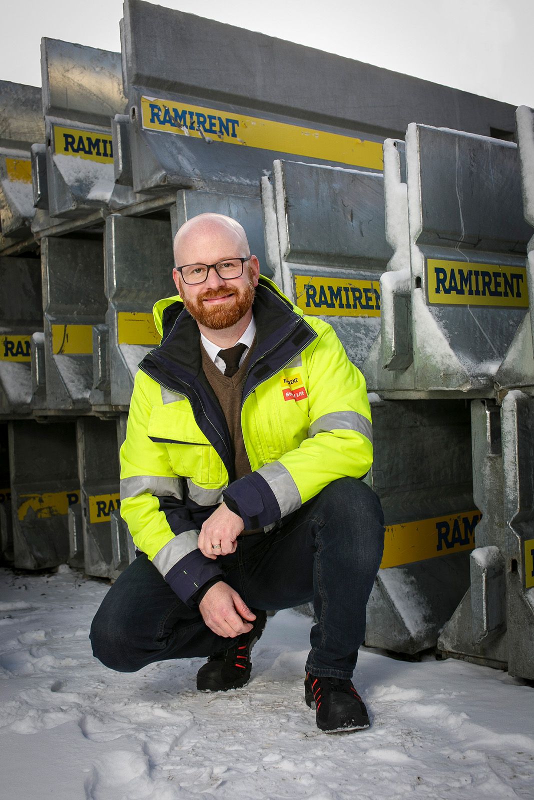 Arbetare i reflekterande säkerhetskläder hukar sig nära Ramirent-utrustning i en snöig industriell miljö