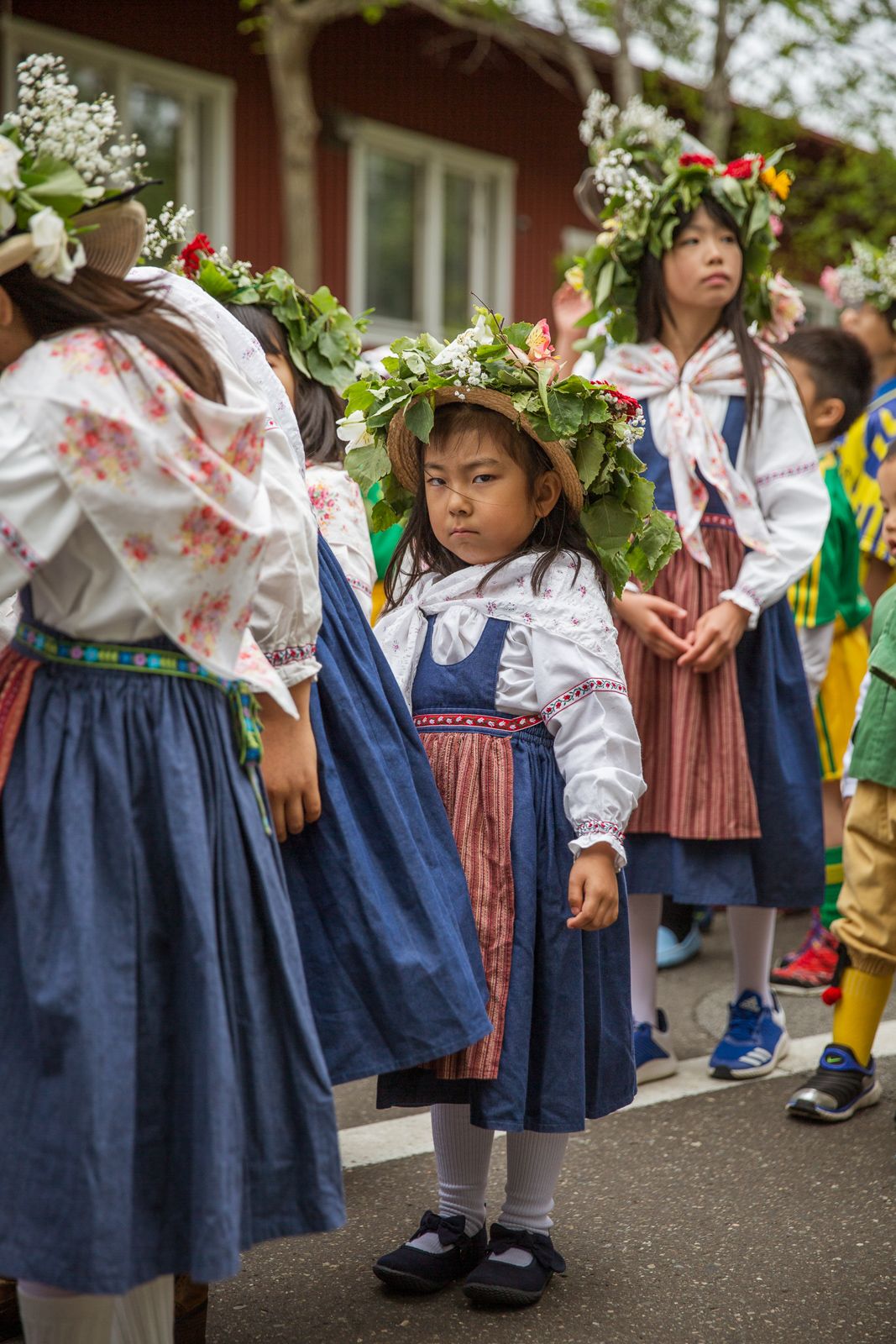 Children wearing traditional blue and white folk costumes with floral wreaths perform at an outdoor celebration.