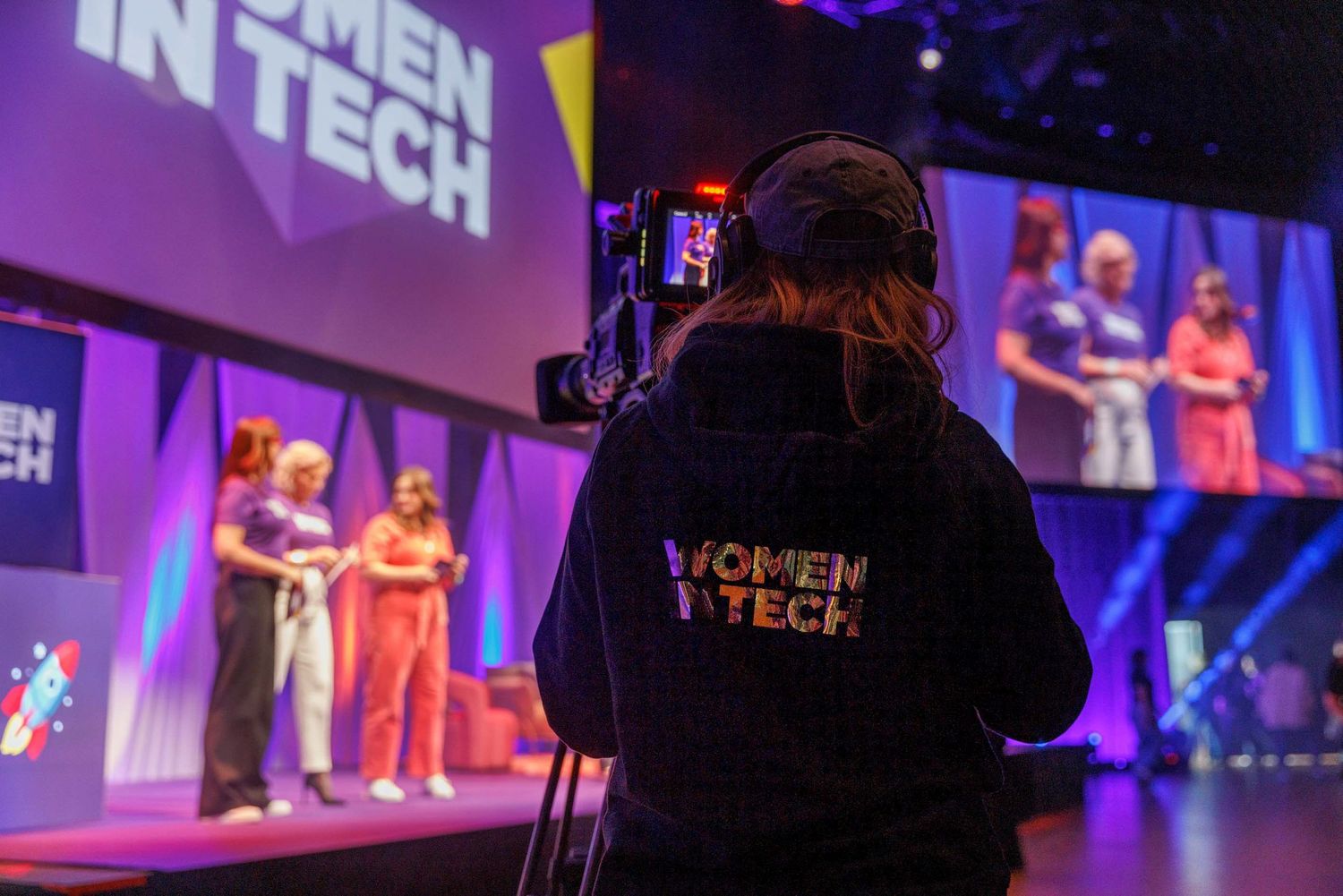 Dark conference hall with Women in Tech branding displayed on large screens.