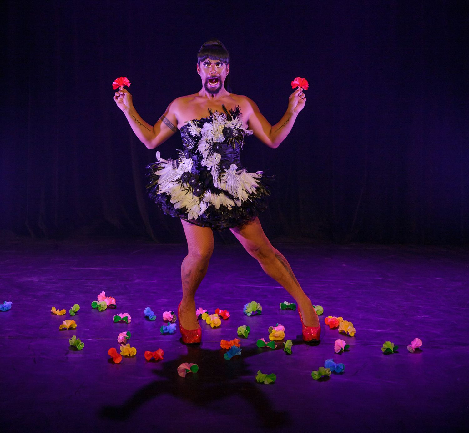 Performer in floral tutu dress dancing among scattered colorful flowers on dark stage.
