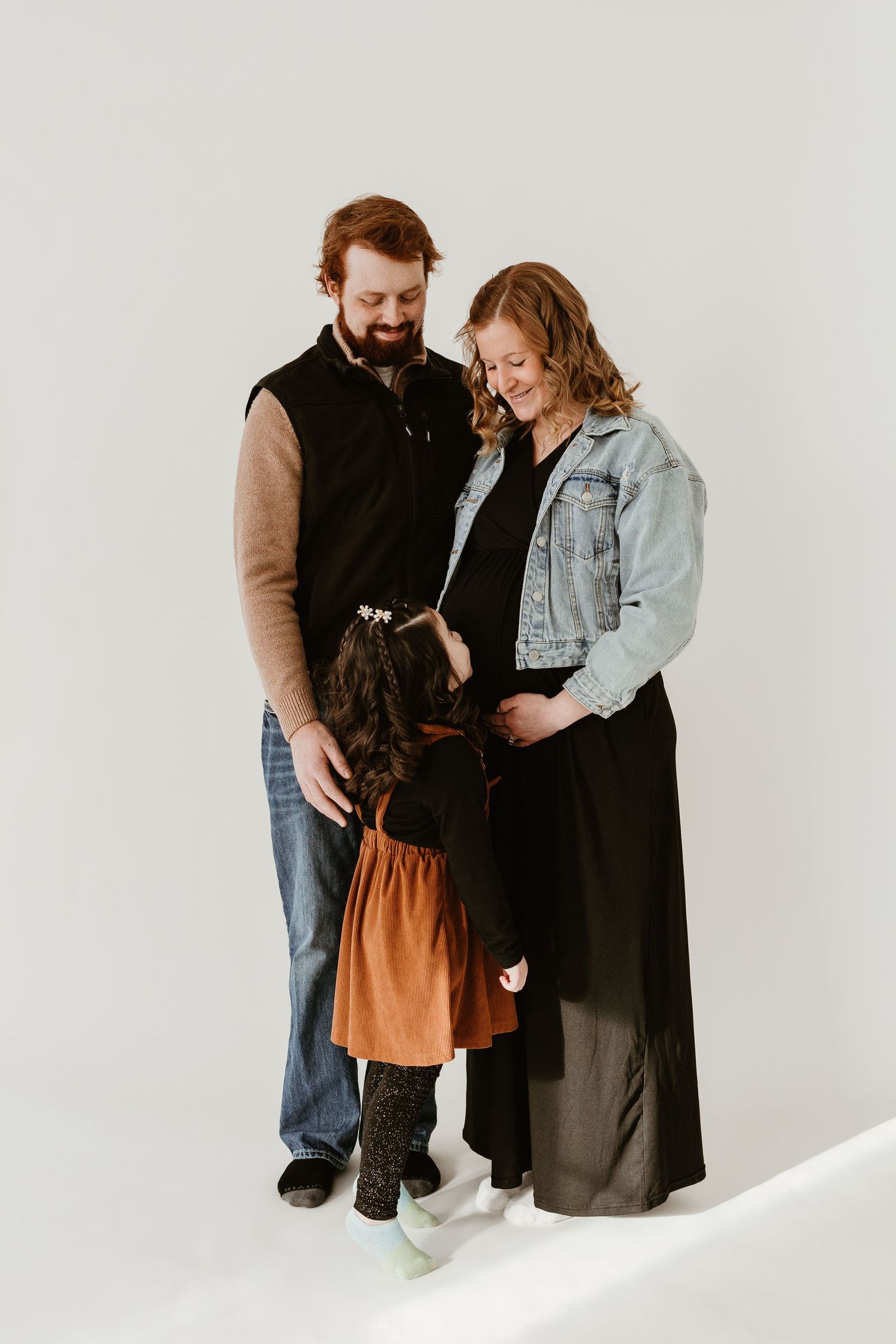 Family maternity photo with red-haired couple and young daughter in rust dress touching mothers pregnant belly.