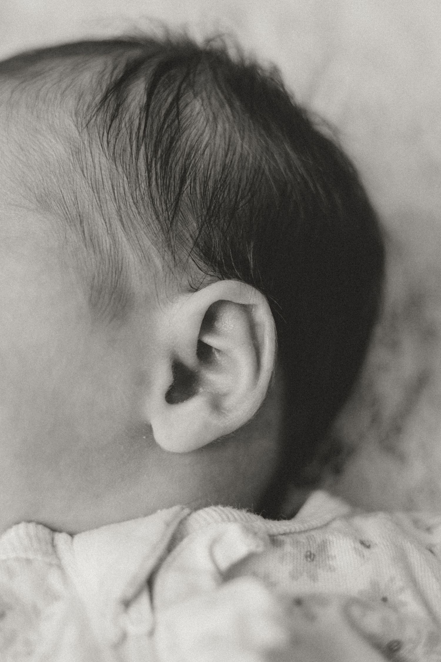 Black and white close-up photo of a newborn baby's ear and dark hair, captured in soft focus.