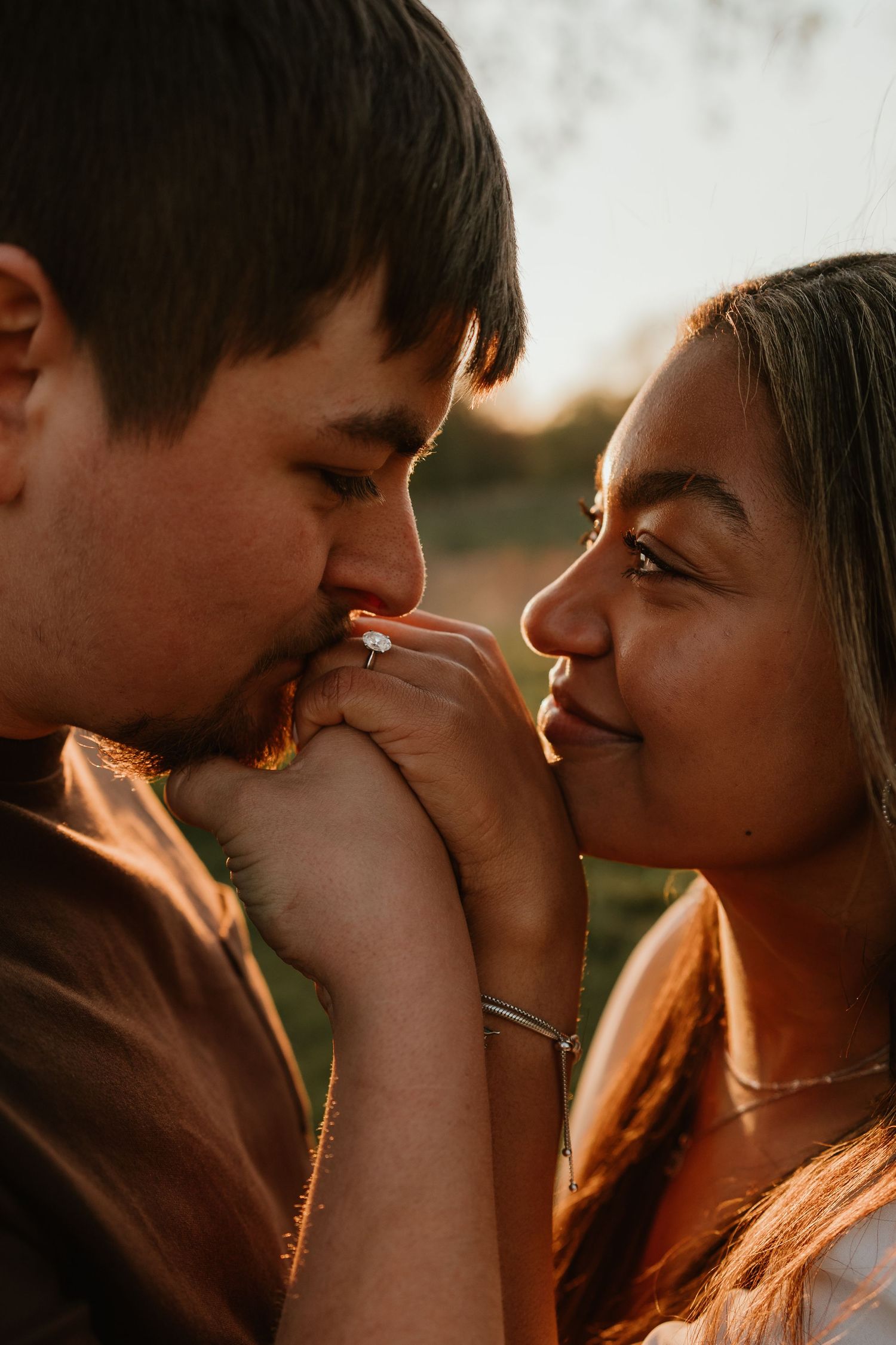 A man kisses a woman's hand adorned with an engagement ring during a romantic golden hour outdoor photo session.