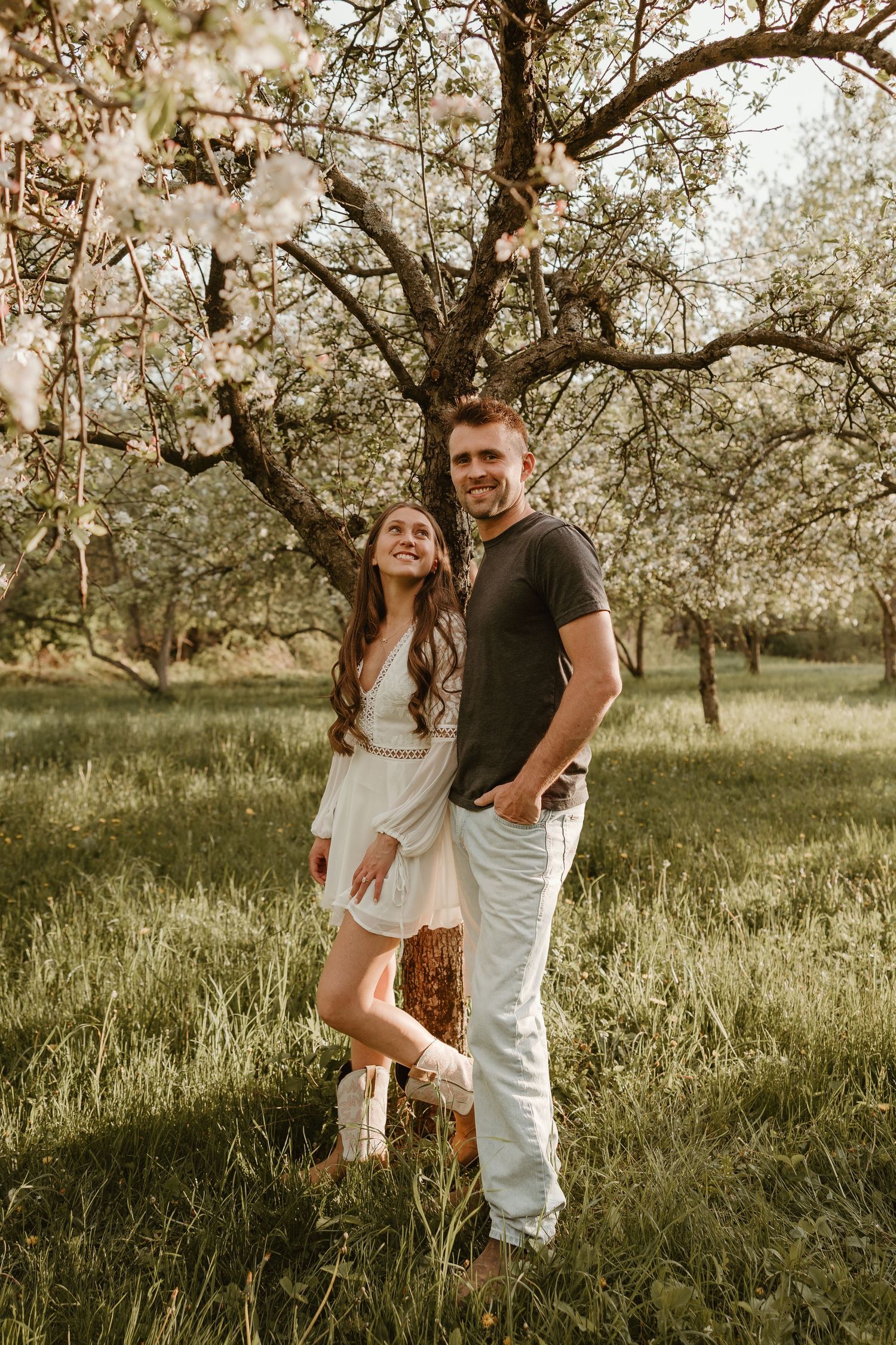 A smiling couple poses together in a blooming spring orchard, woman in white dress and man in gray tee.
