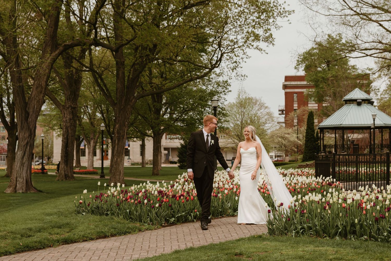Bride and groom walk hand-in-hand along a brick path surrounded by colorful tulips and lush trees at an outdoor wedding venue.