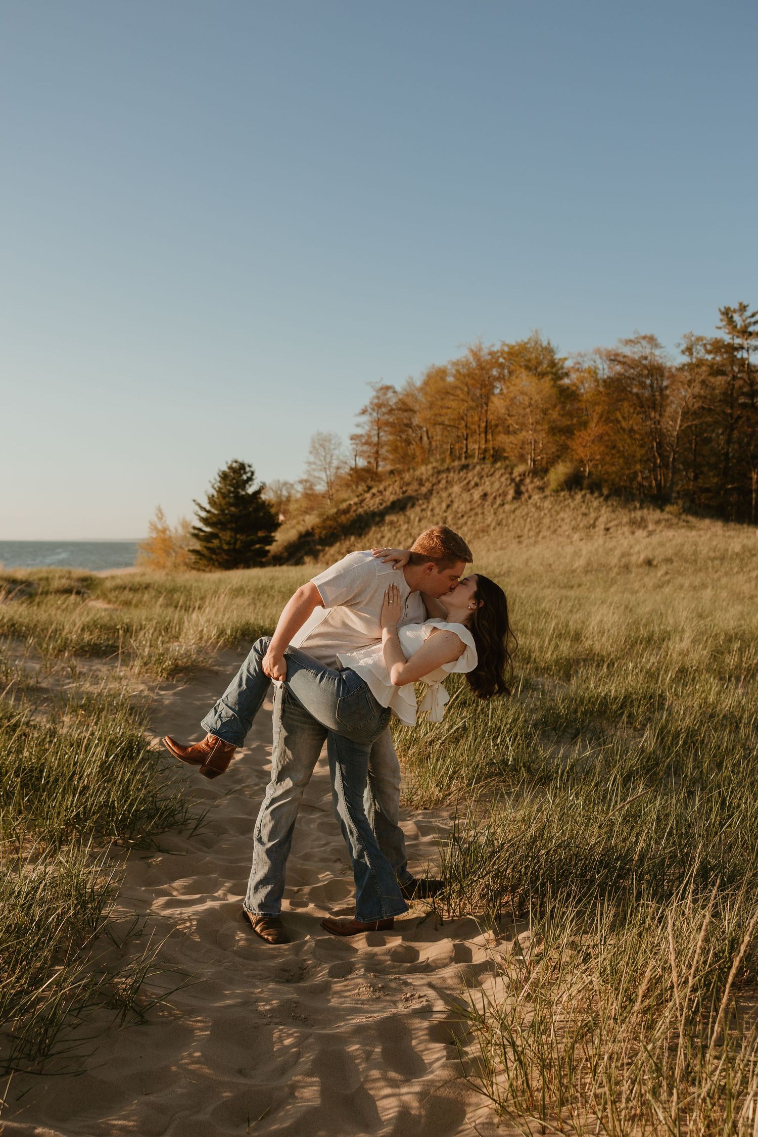 Couple sharing a romantic kiss on sandy beach dunes with golden autumn trees and blue sky in background.