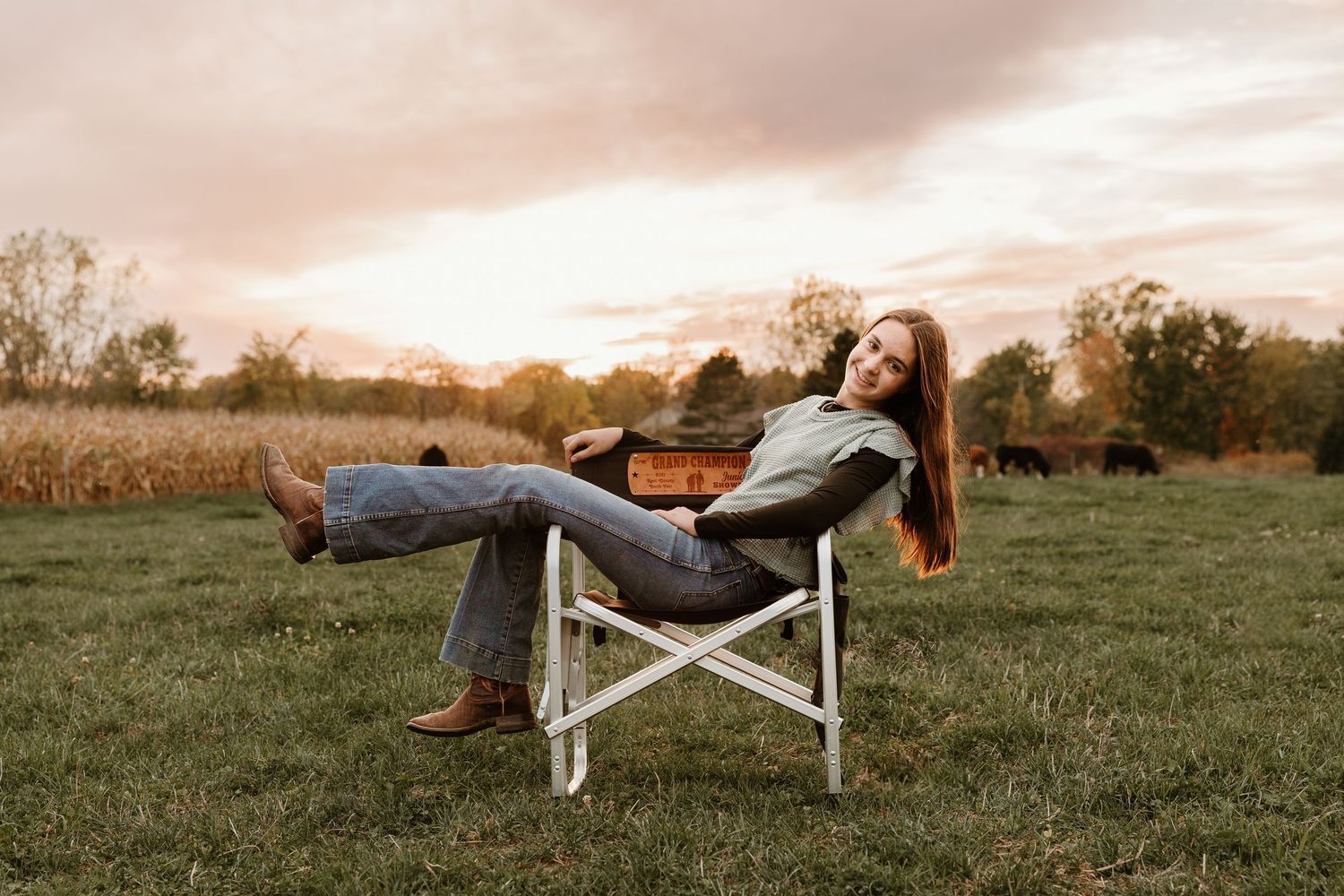 Woman relaxing in a folding chair on a green field at sunset, wearing jeans and brown boots with cattle in background.