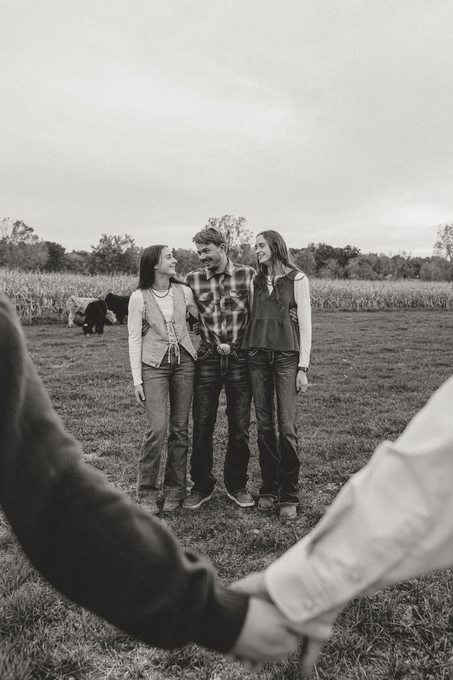 Black and white photo of three friends laughing in a farm field, holding hands in a circle outdoors.