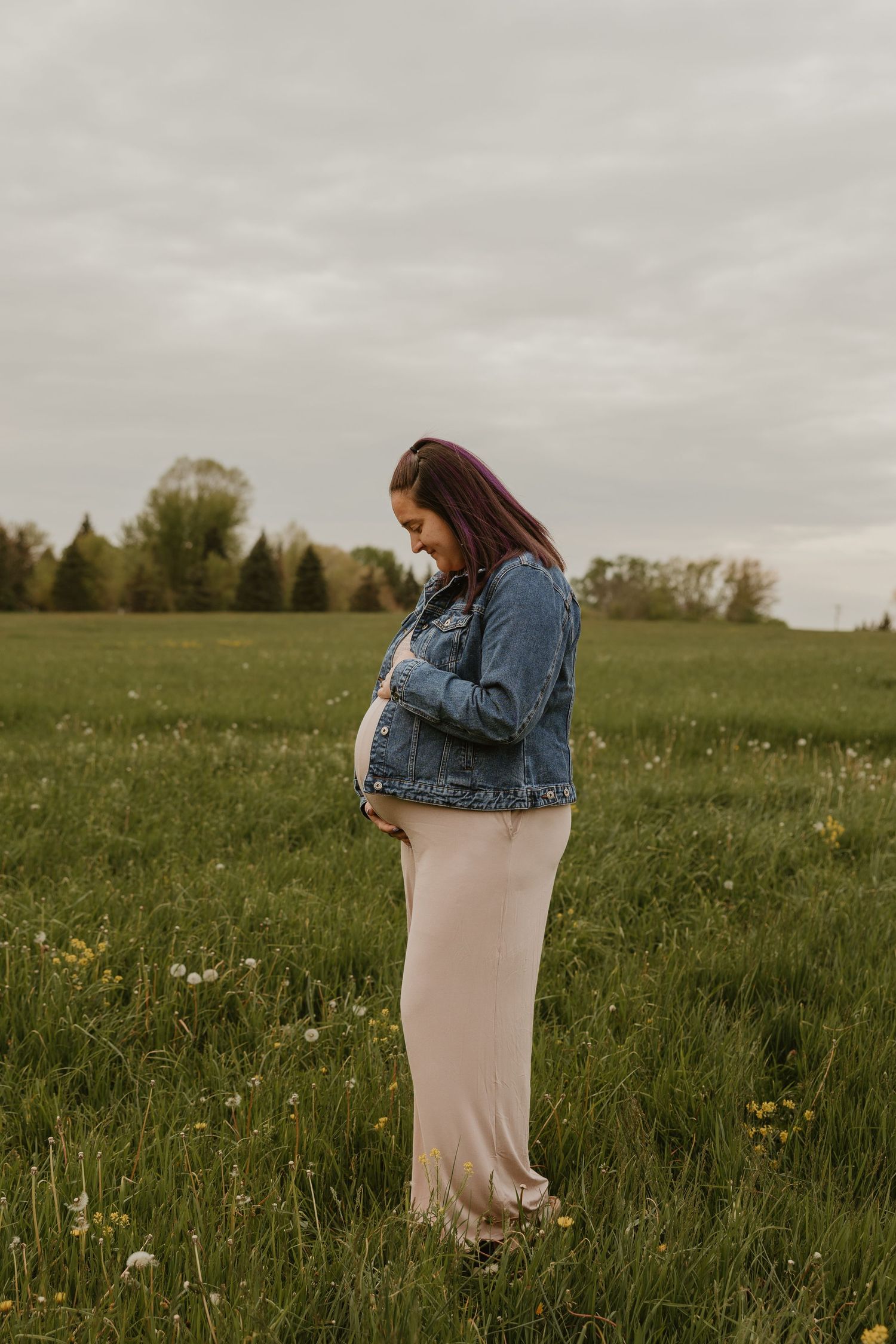 Pregnant woman in denim jacket and blush maxi dress standing in green field, looking down at bump.