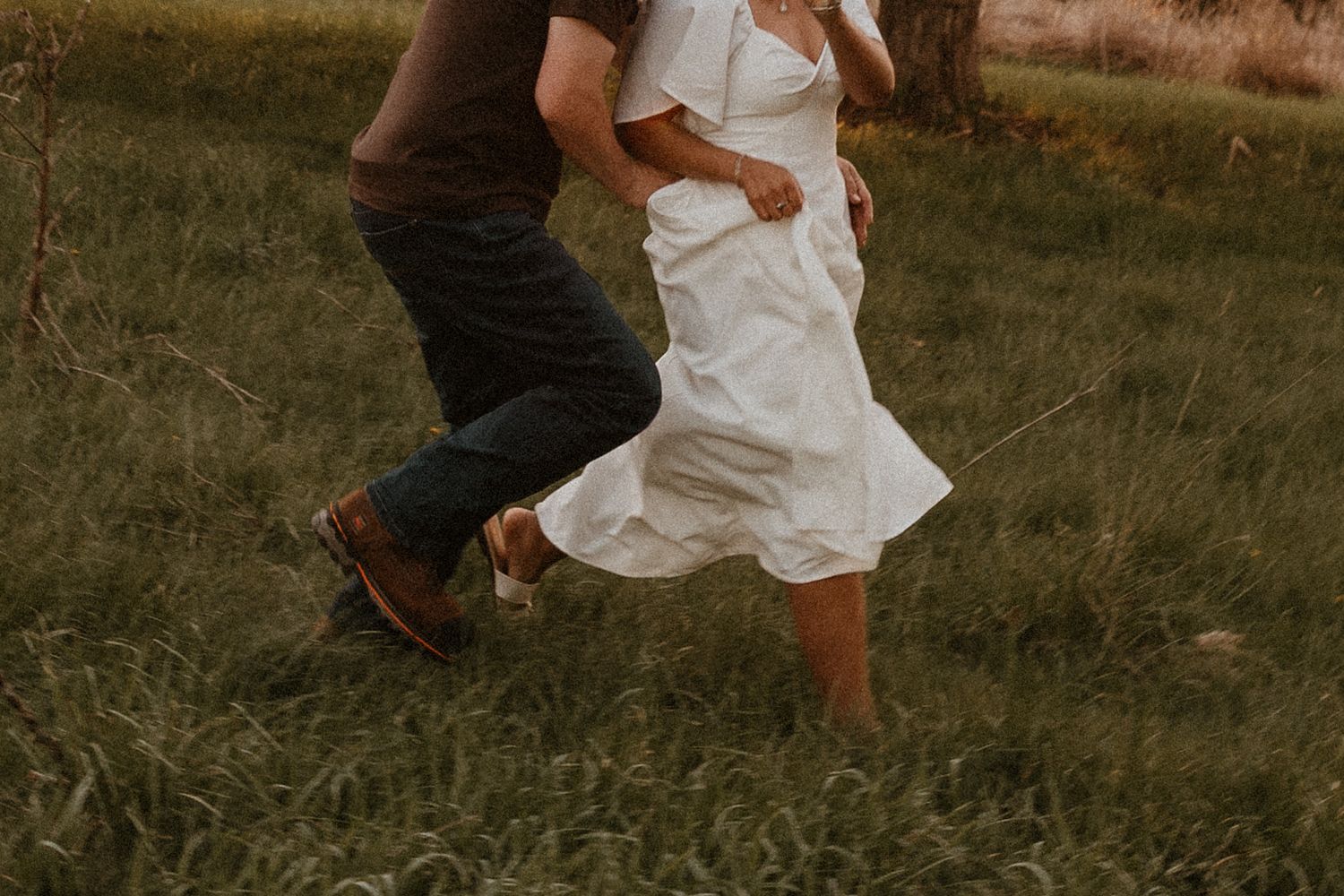 Couple running through a green grassy field, woman in white dress and man in jeans and brown boots.