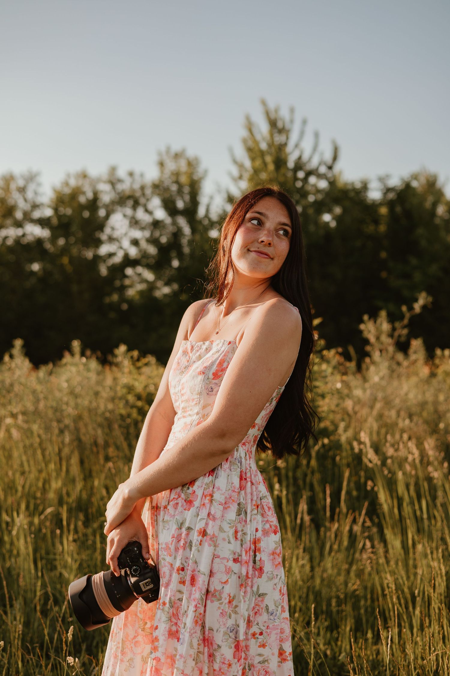 Young woman in floral dress holding a camera, standing in a sunlit field with green trees in the background.