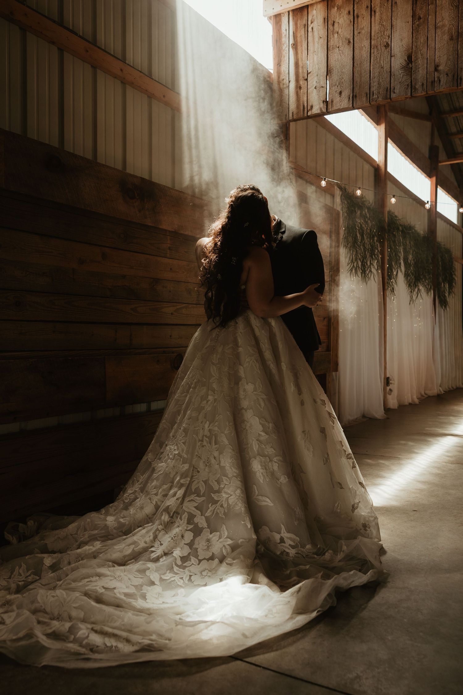 A bride in a lace ballgown with a long train stands embracing her groom in a rustic barn with sunlight streaming in.