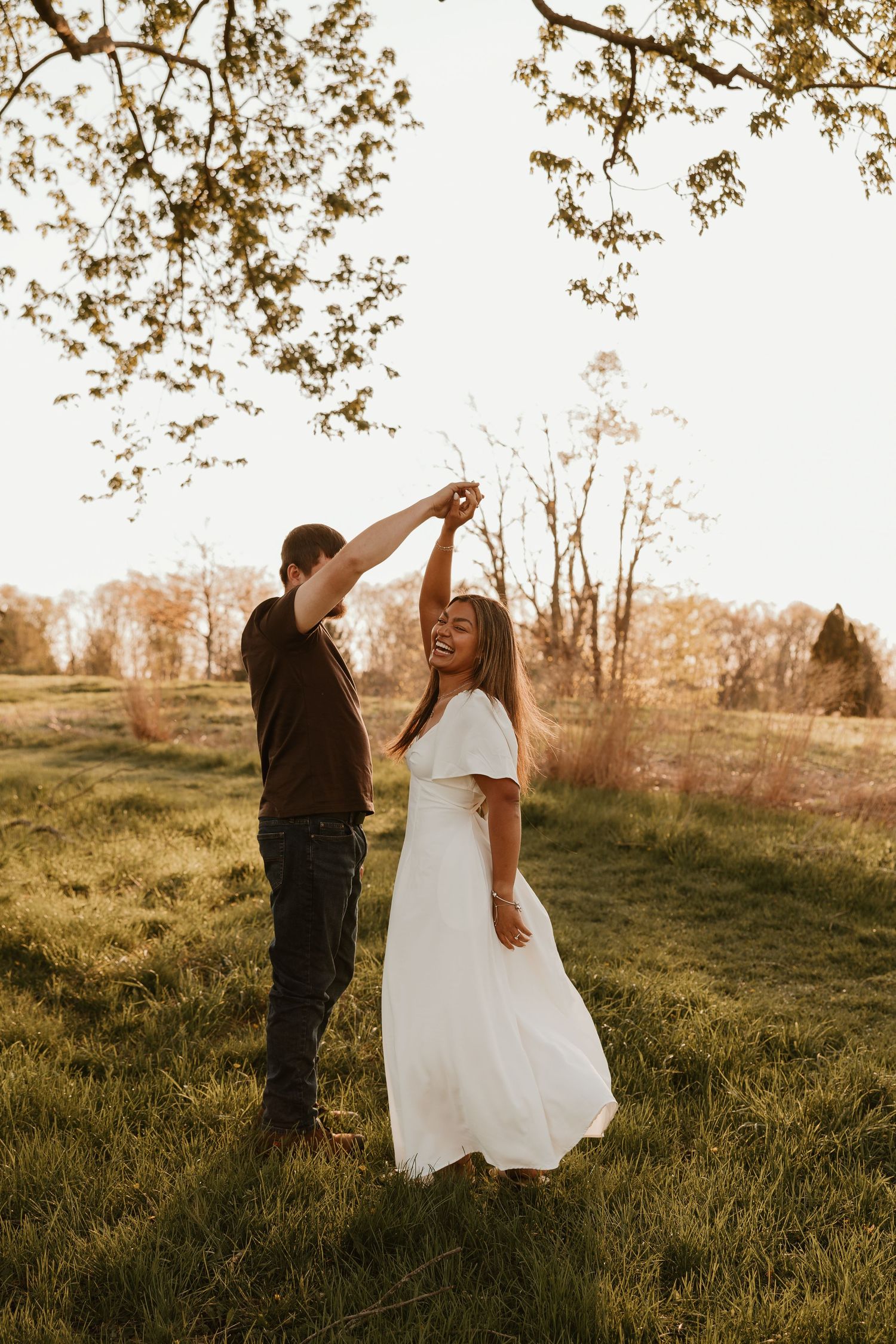Couple dancing in a sunlit field, woman in white dress twirling under a tree at golden hour.