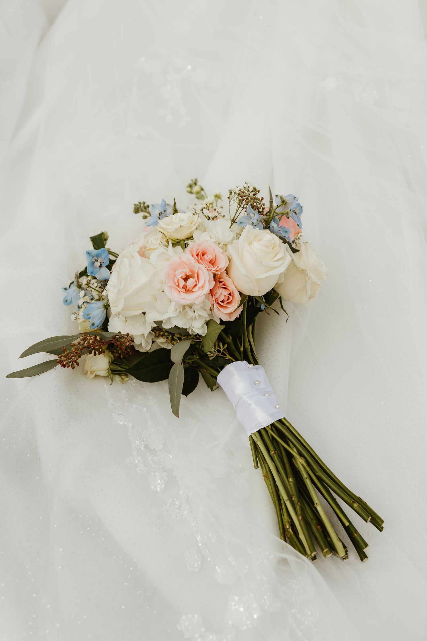 A bridal bouquet of white roses, pink blooms, and blue flowers tied with white ribbon on a wedding dress.