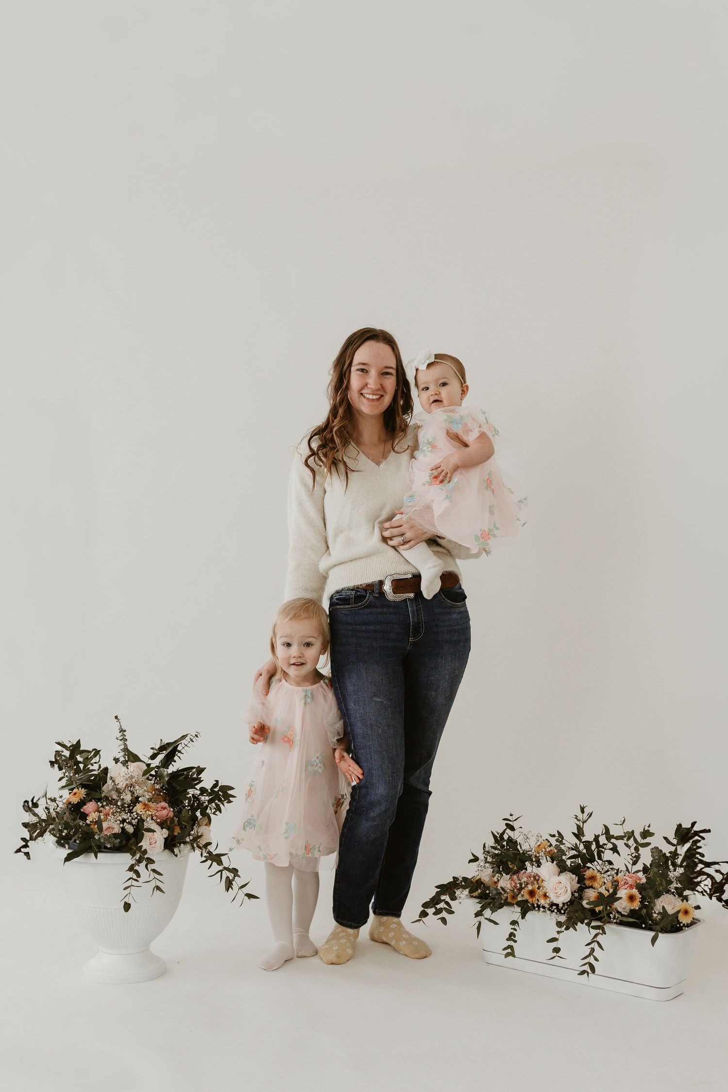 A smiling mother holds a baby in pink while a toddler stands beside her, surrounded by floral arrangements.