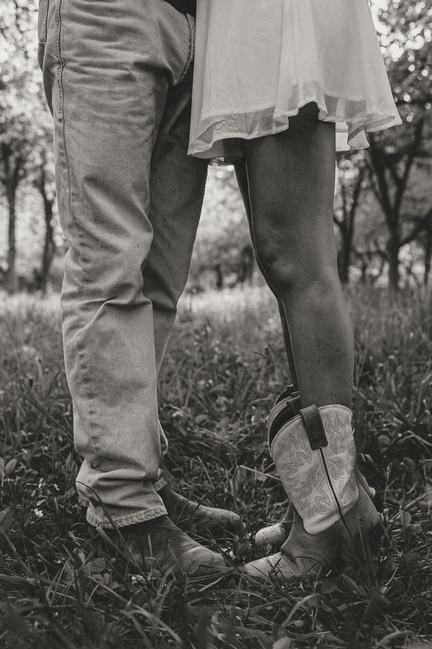 Black and white photo of couple standing close in grass, woman wearing cowboy boots and white dress.