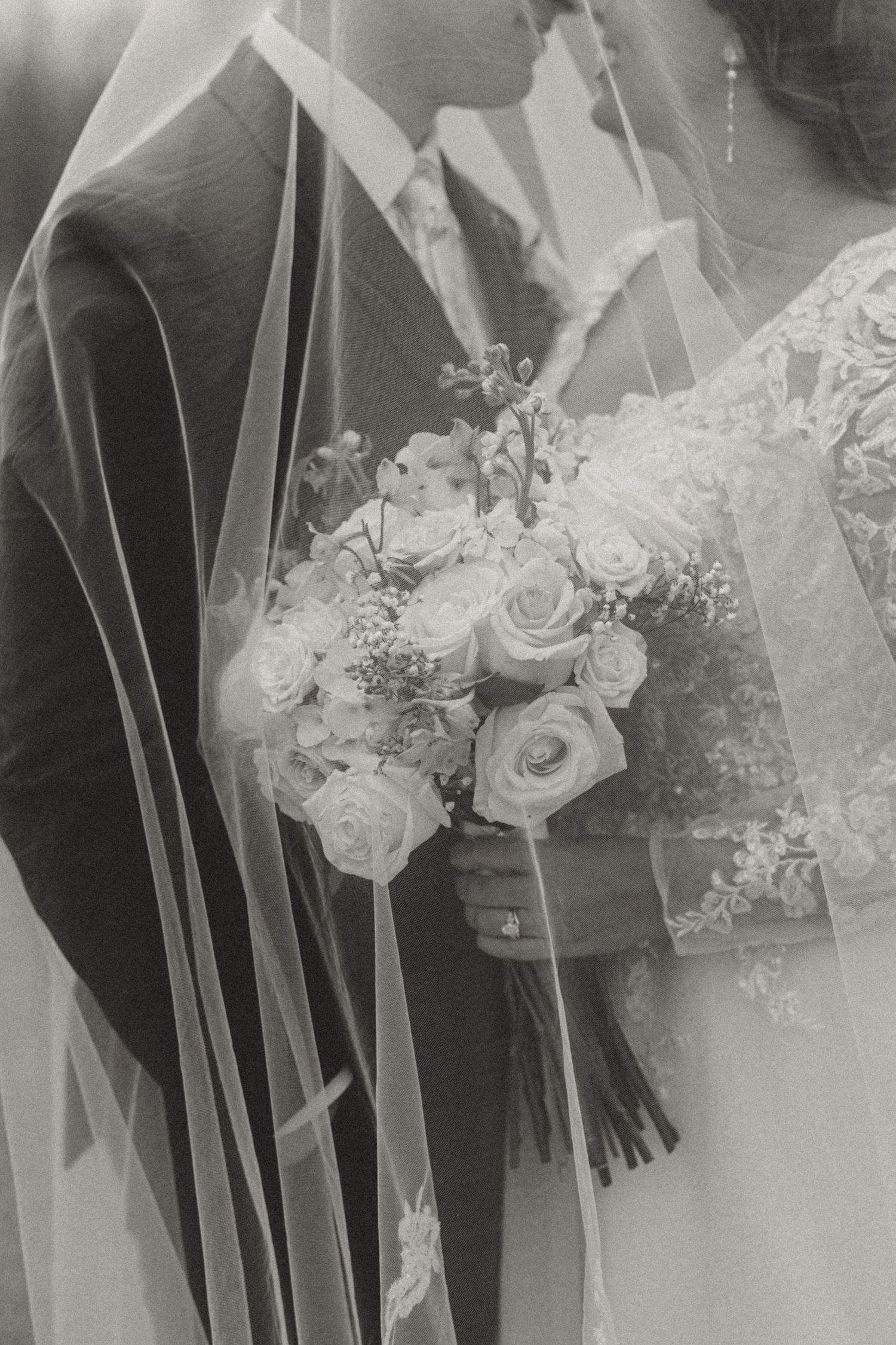 Black and white wedding photo of bride holding rose bouquet under veil beside groom in suit.