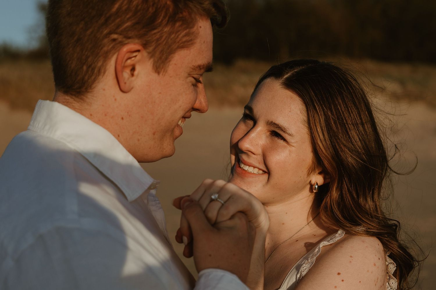 Couple sharing a romantic moment outdoors, woman smiling with engagement ring visible, golden hour lighting.