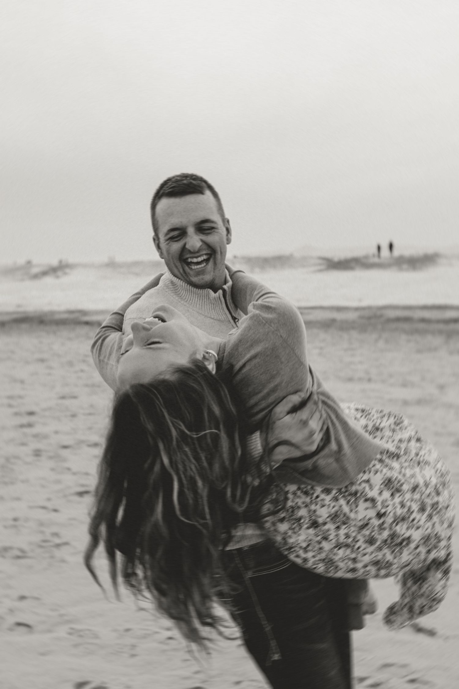 A joyful couple laughs and embraces on a beach, black and white photo capturing a romantic playful moment.