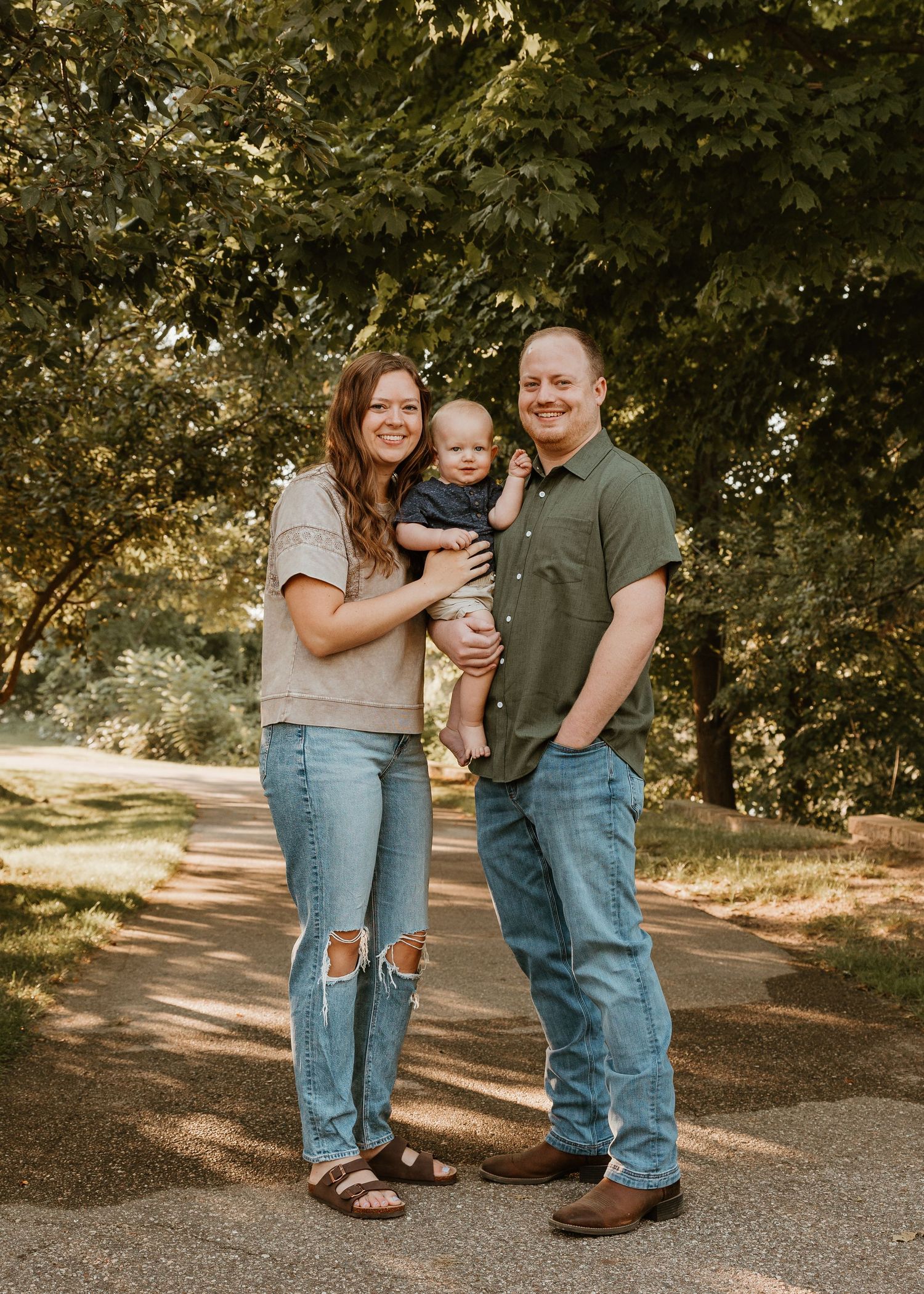 A smiling family of three poses outdoors on a tree-lined path, with parents holding their baby in a sunny park.