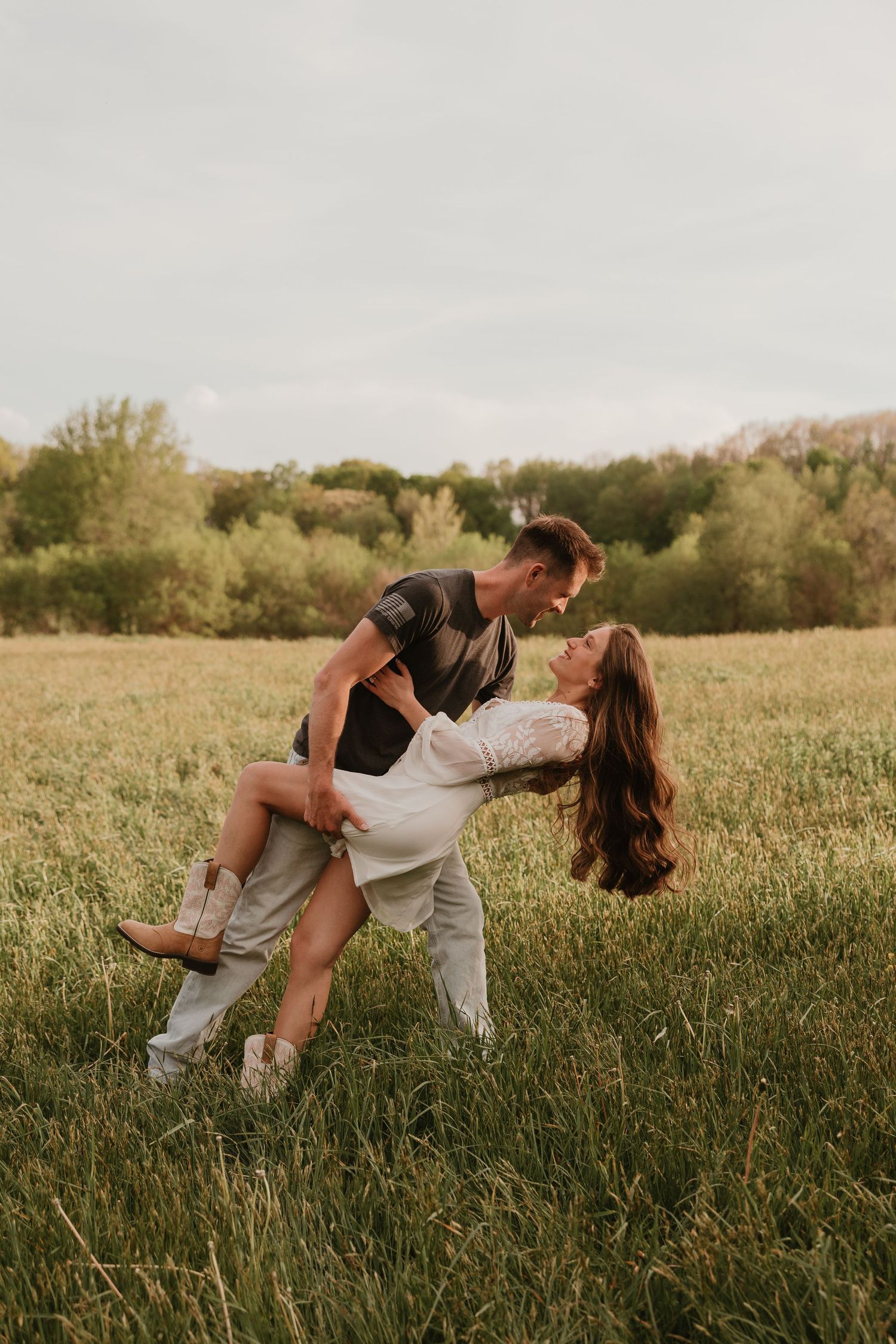 Couple dancing in a green field, man dipping woman in white dress and cowboy boots during romantic outdoor photo session.