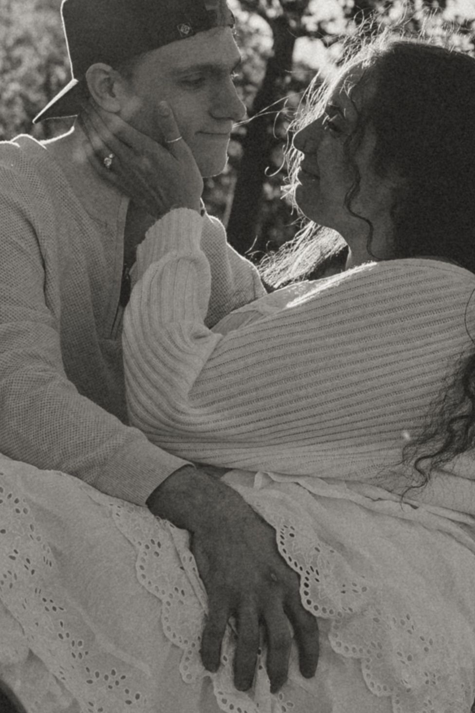 Black and white photo of a romantic couple embracing outdoors, woman tenderly touching mans face in soft natural light.