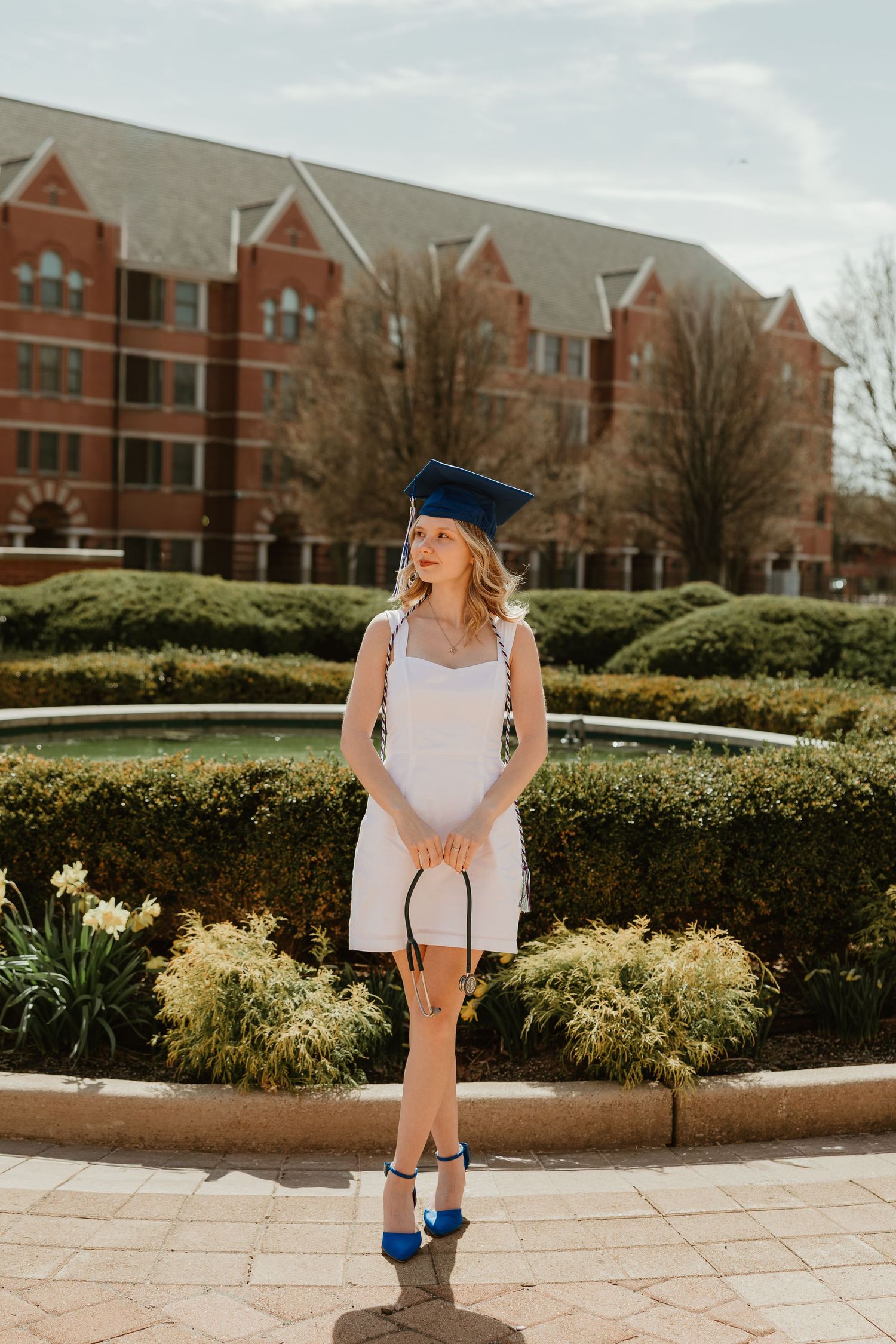 Young woman in white dress and blue graduation cap poses by fountain with brick university building backdrop.