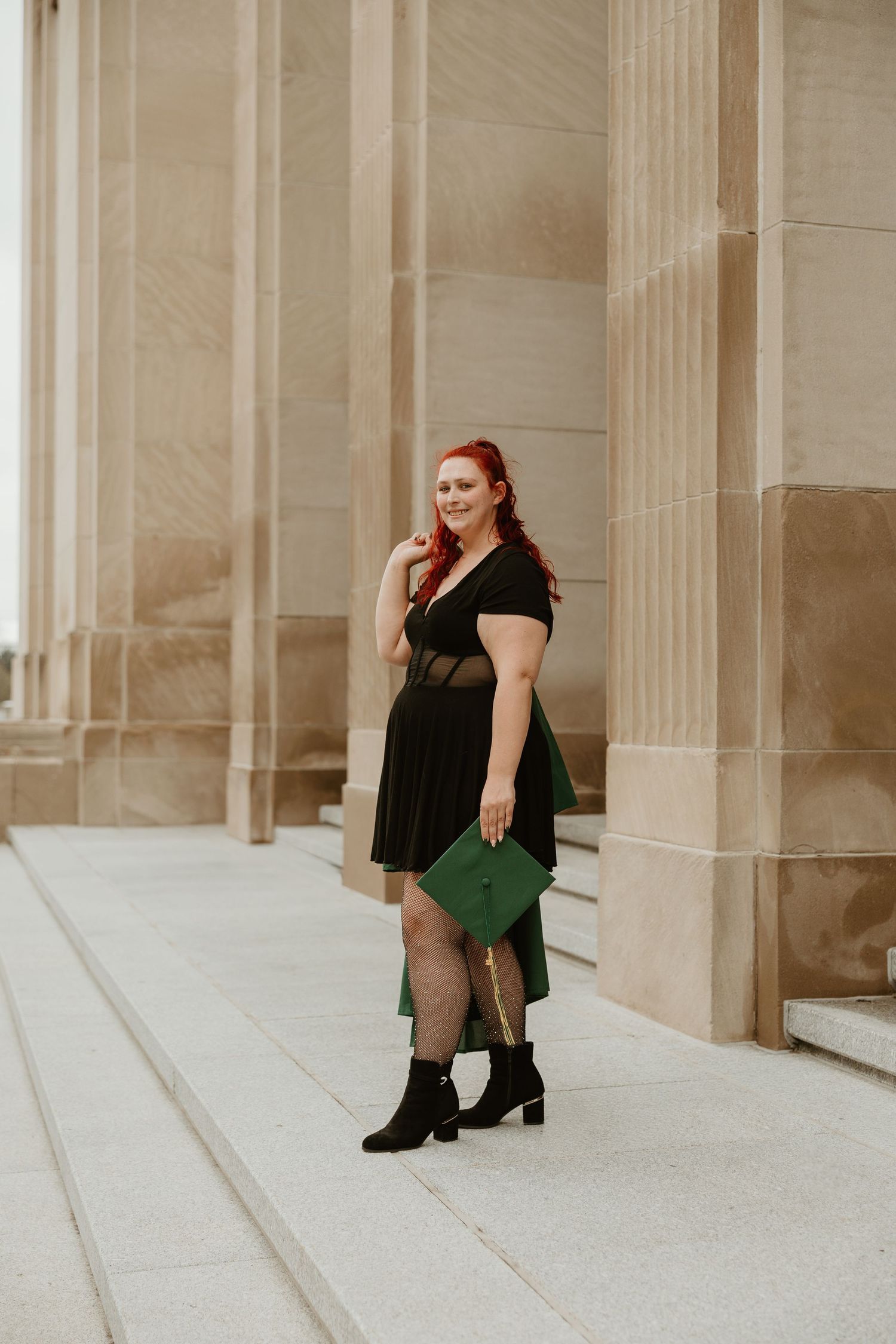 A woman with red hair in a black dress holds a green graduation cap while posing between stone columns.