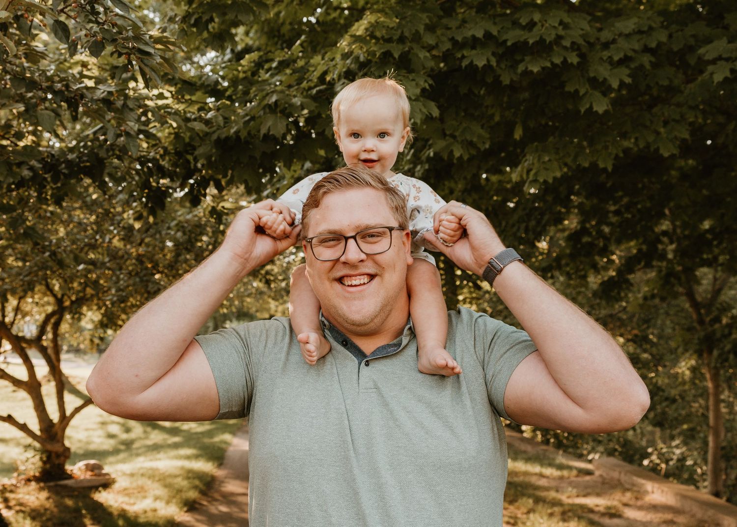 Man with glasses smiling outdoors holding a toddler on his shoulders among green trees.