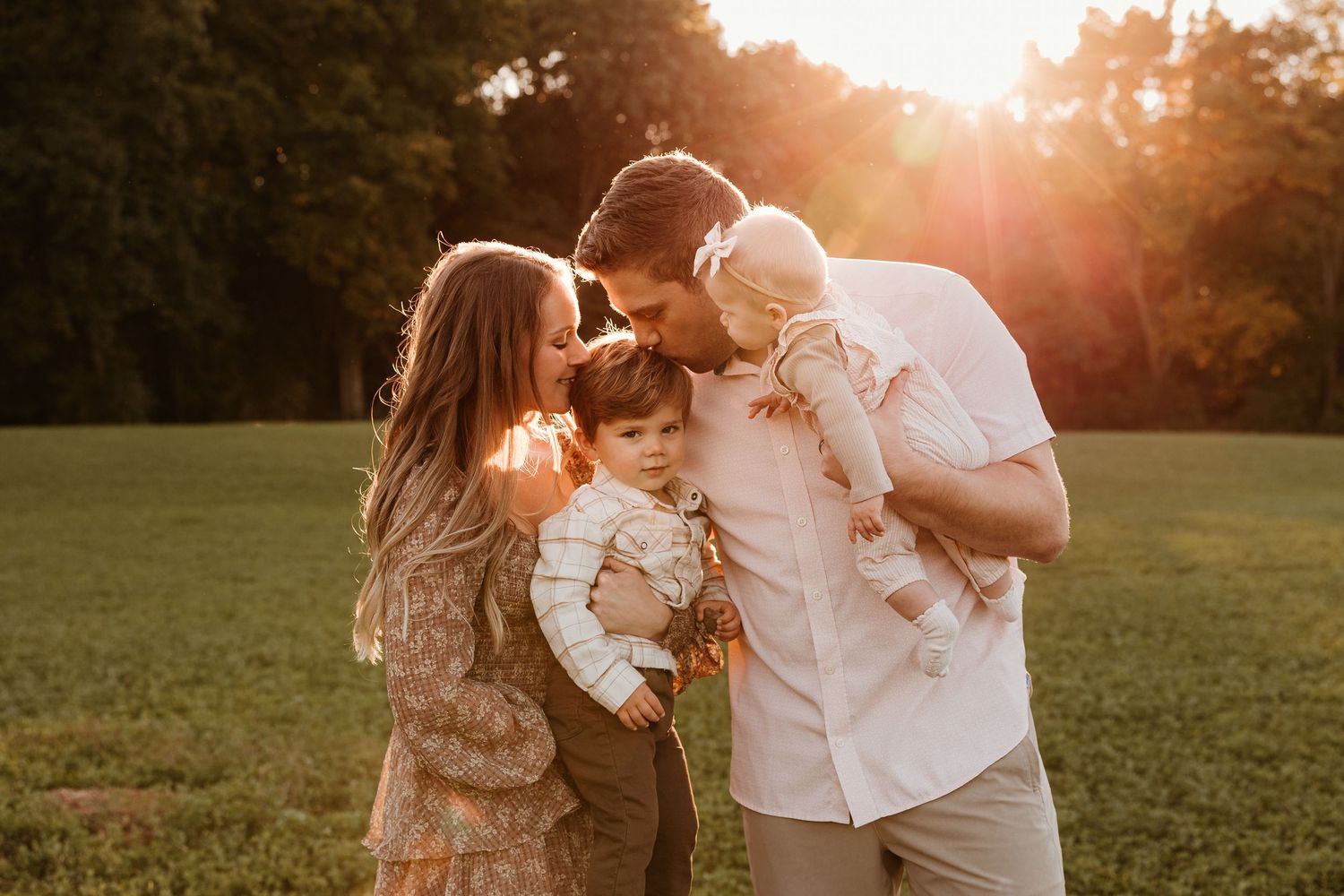 Family portrait at golden hour with parents kissing toddler and baby in a sunlit green field.