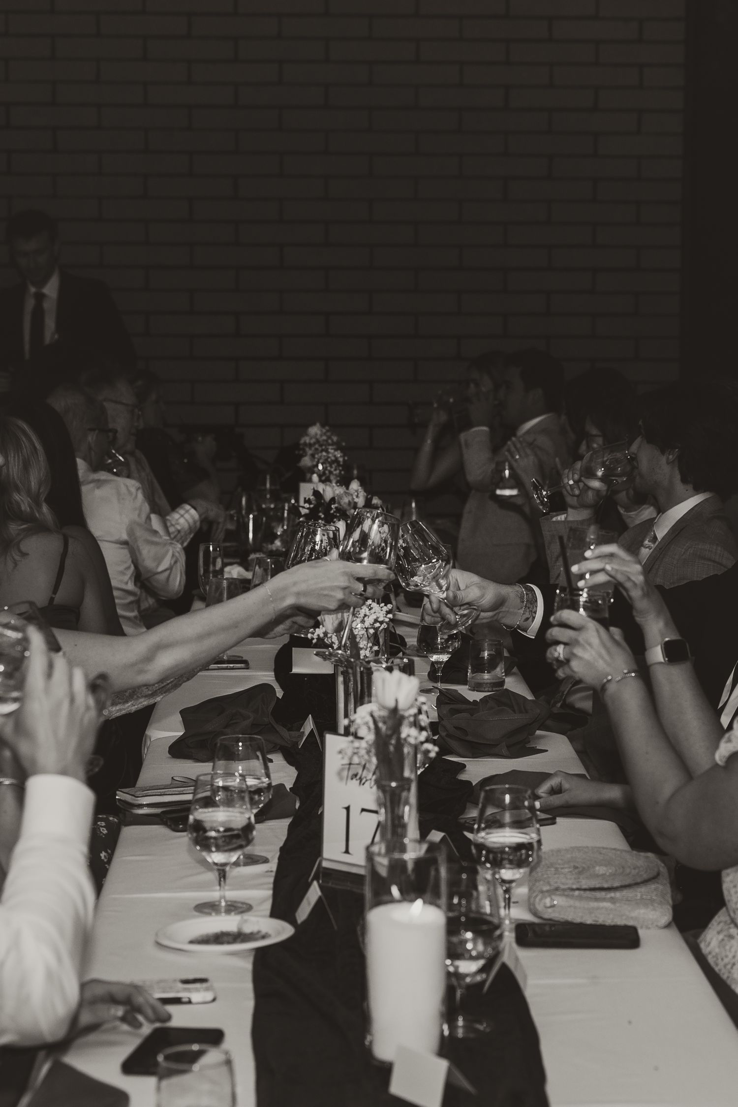 Black and white photo of wedding guests raising glasses in a toast at a long decorated reception table with candles.