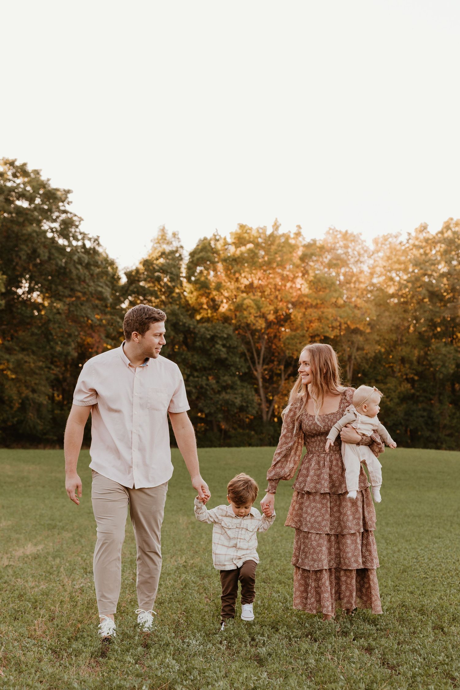 Family of four walking in a sunlit green field at golden hour, parents holding toddler's hands while mom carries baby.