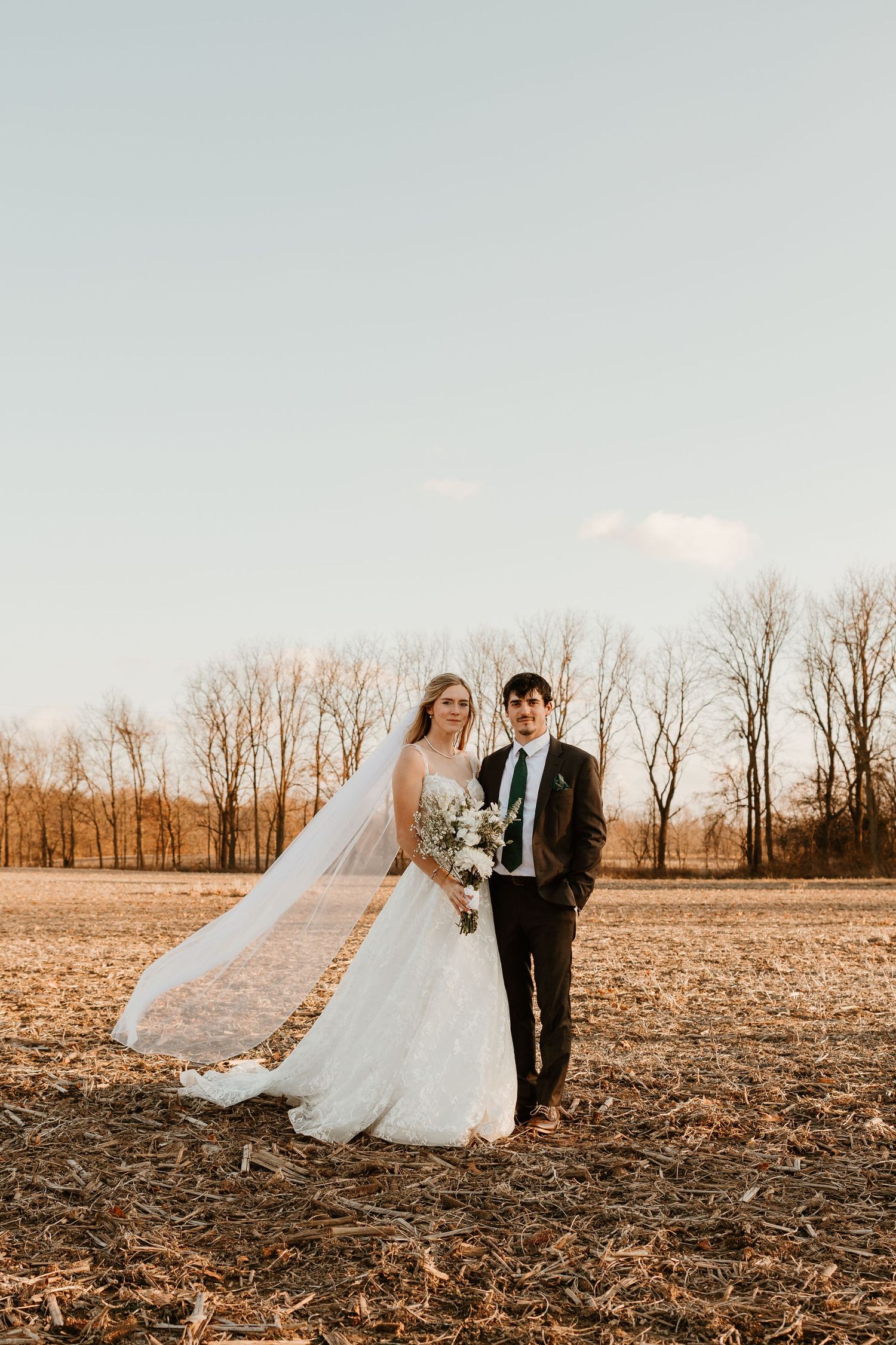 Bride and groom pose in a golden field at sunset, her veil flowing in the wind, with bare trees in the background.