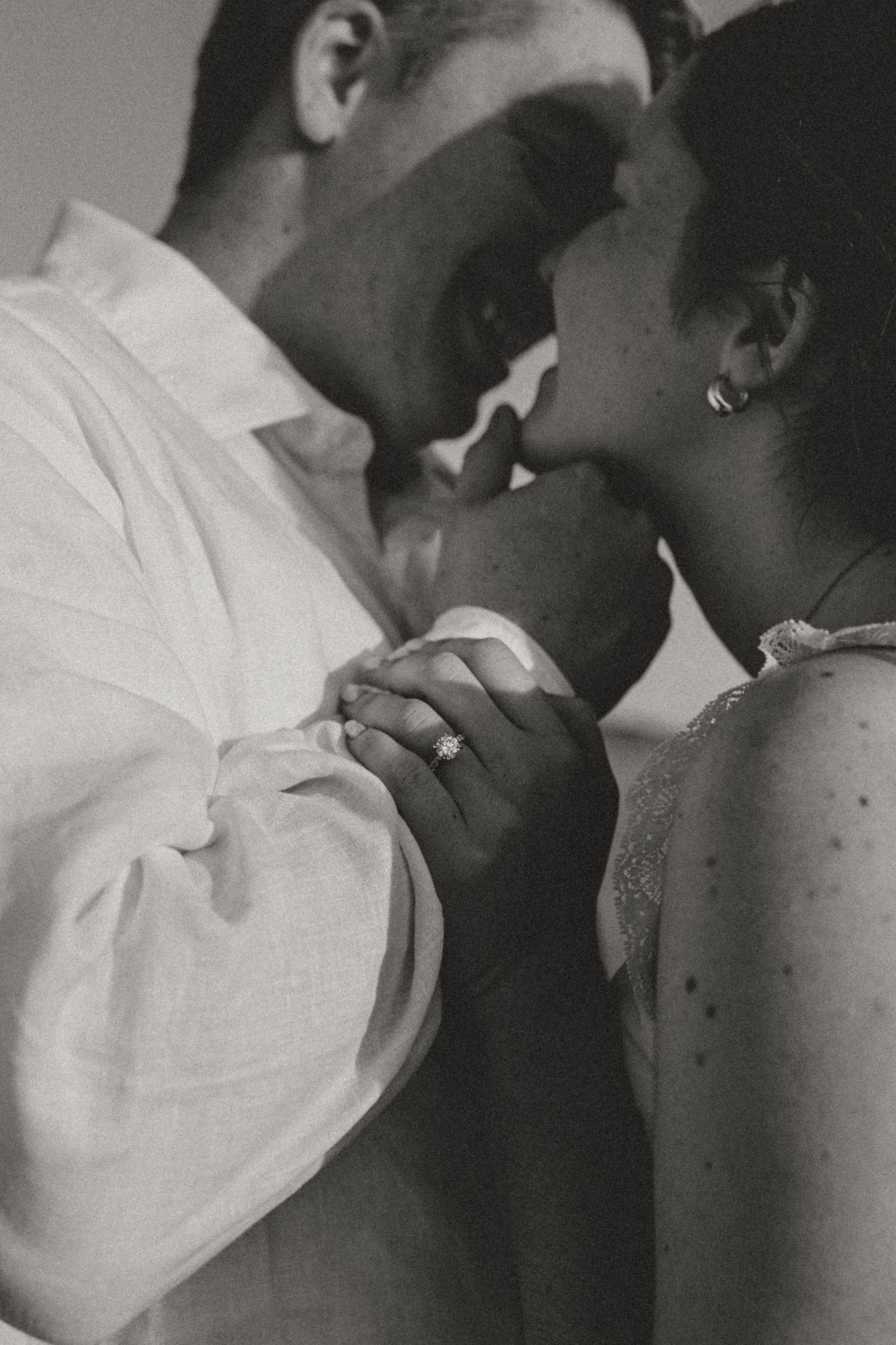 Black and white photo of a couple sharing a romantic kiss, holding hands with an engagement ring visible.
