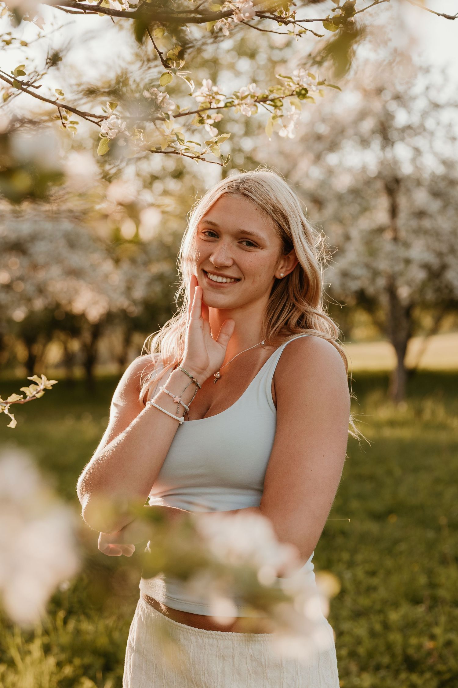Smiling blonde woman in white crop top posing near blooming spring trees in golden hour sunlight.