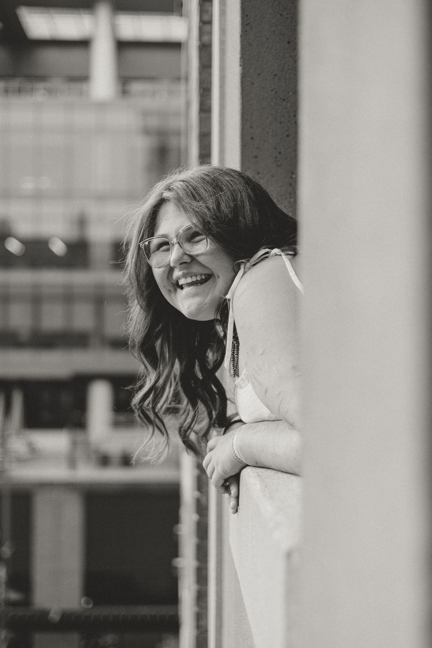 Black and white portrait of a smiling woman with glasses and long wavy hair leaning against a pillar outdoors.