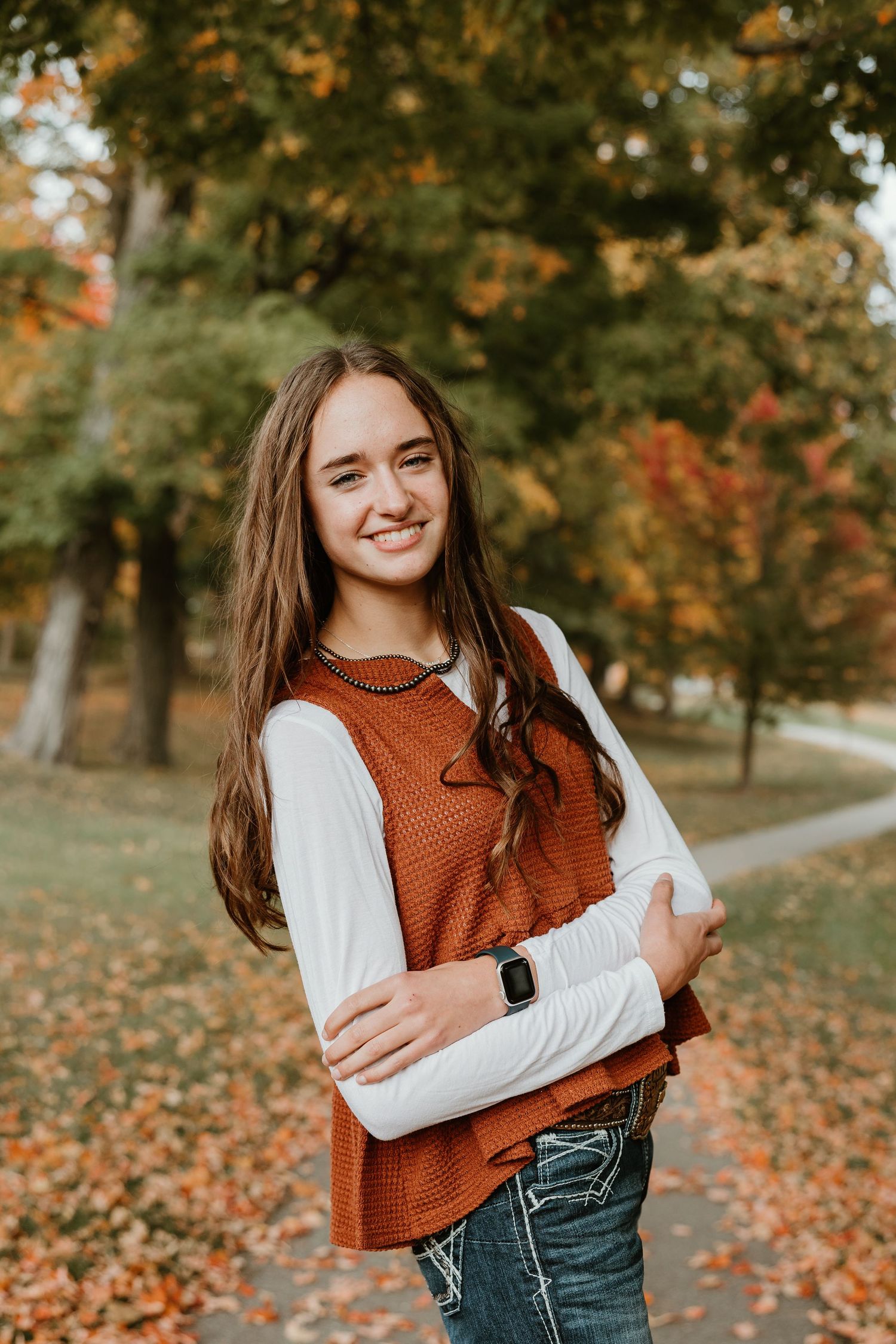 Smiling teenage girl in rust orange vest and white long-sleeve shirt poses outdoors among colorful fall foliage.