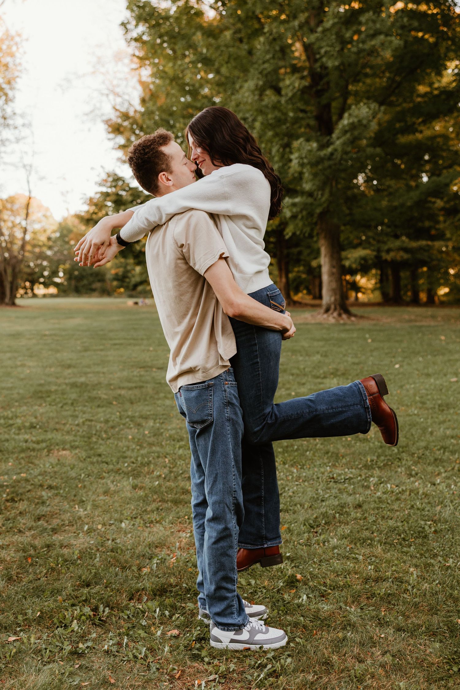 Couple sharing a romantic kiss in a park, man lifting woman in jeans and casual tops among green trees.