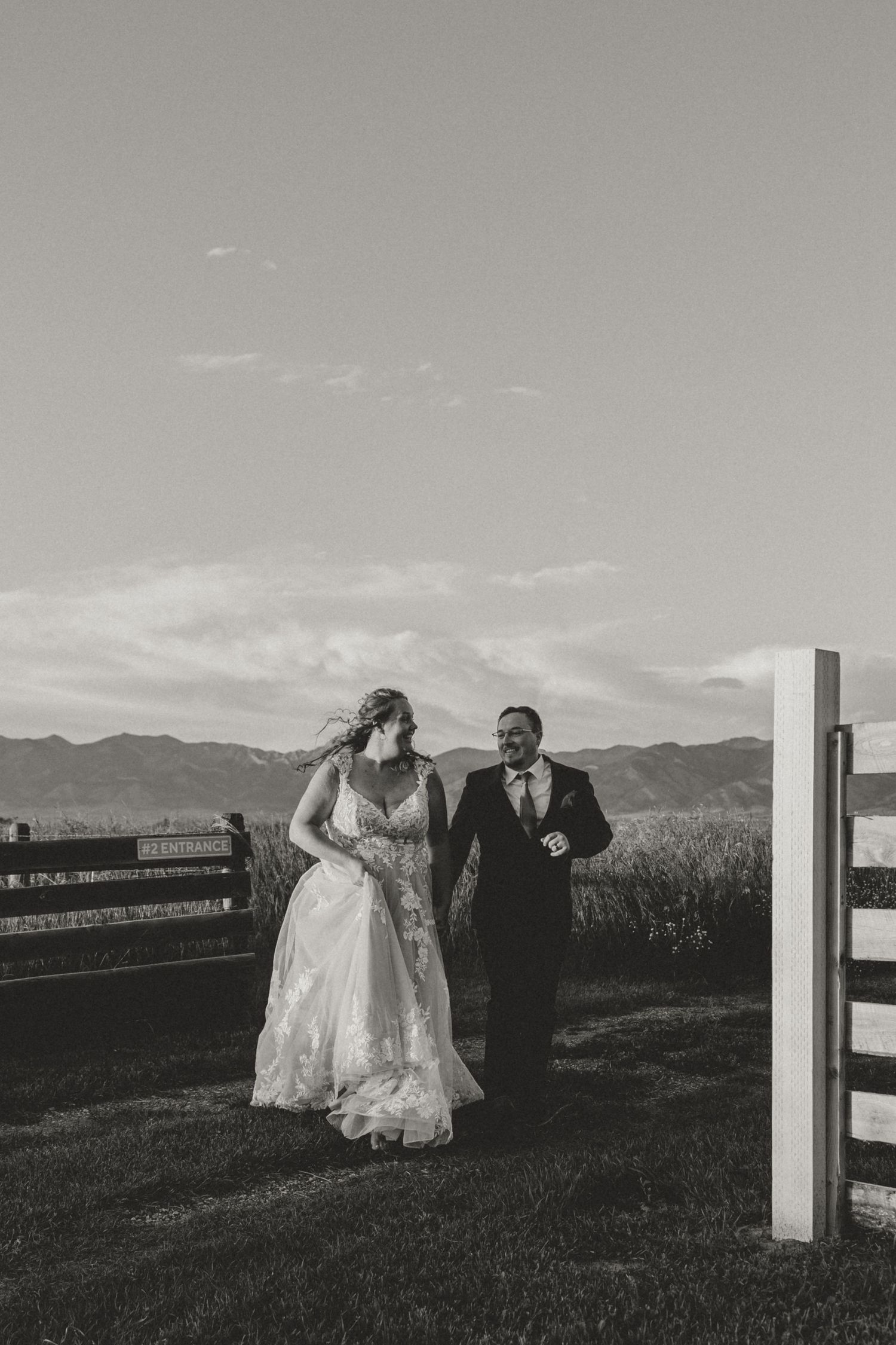 Black and white photo of a bride and groom walking hand in hand by a fence with mountains in the background.
