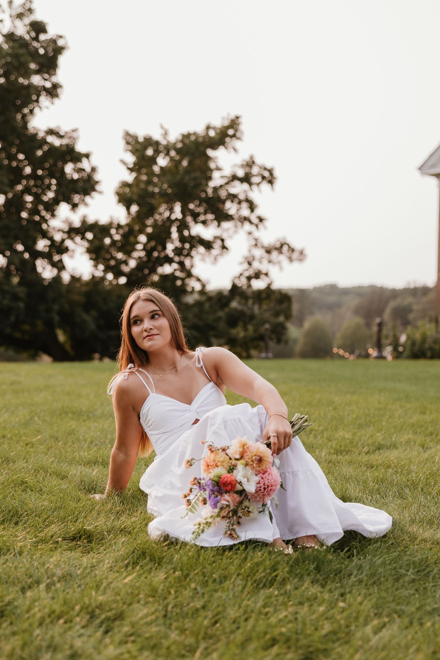 Bride in white dress sitting on green grass holding colorful bouquet of flowers outdoors at dusk.
