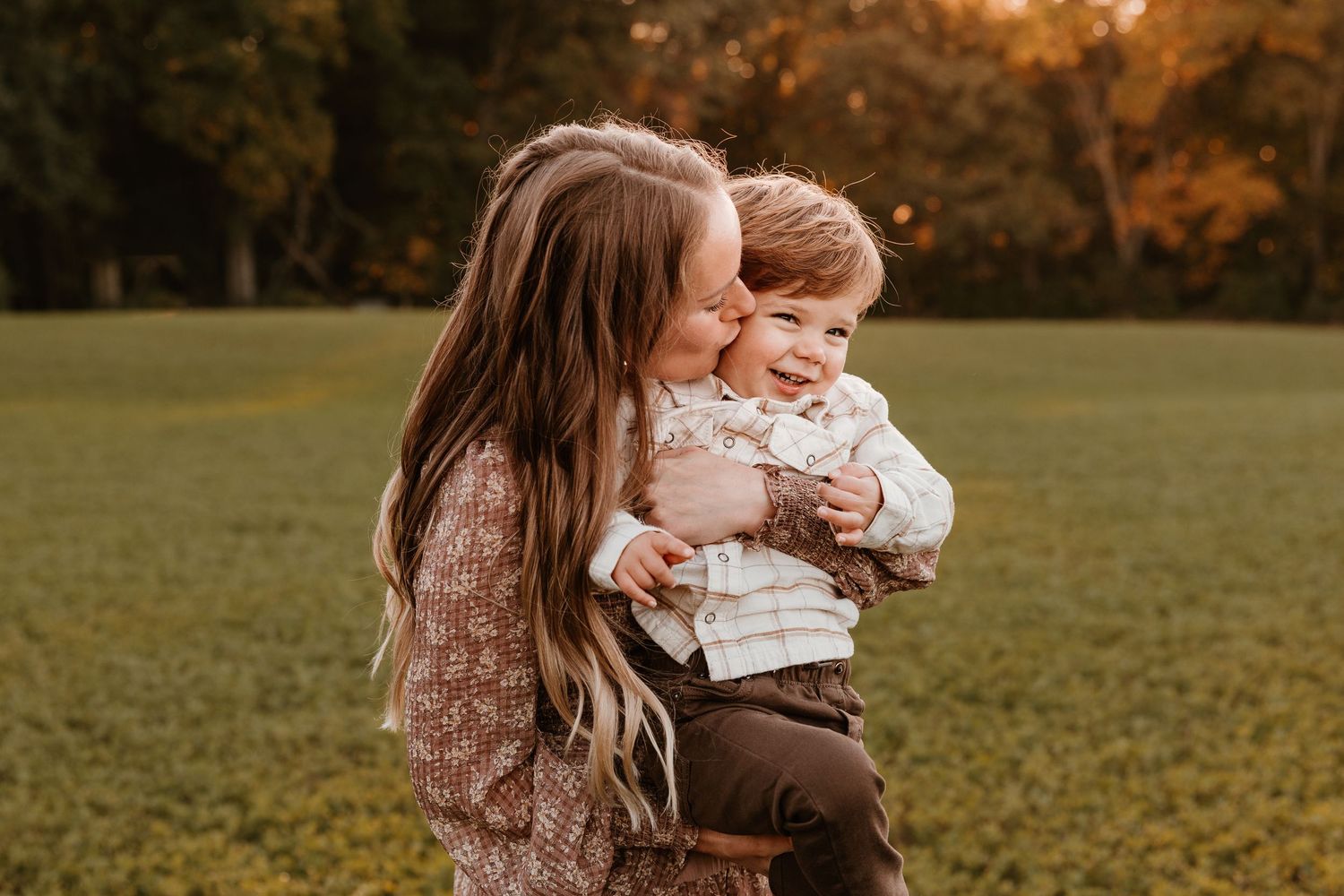 A mother kisses her smiling toddler son outdoors in a golden autumn park setting.