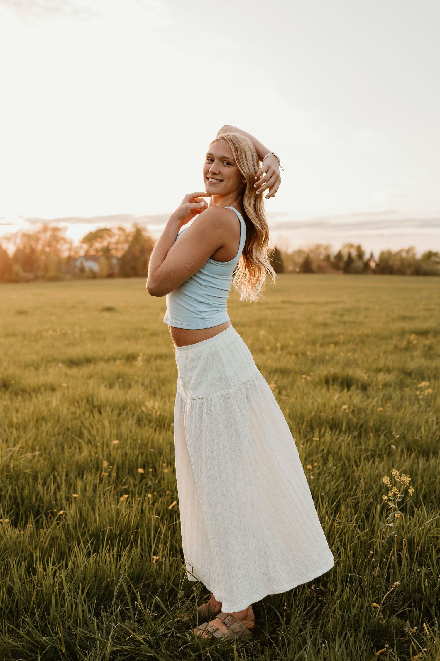 Smiling blonde woman in light blue crop top and white maxi skirt standing in green field at golden hour sunset.