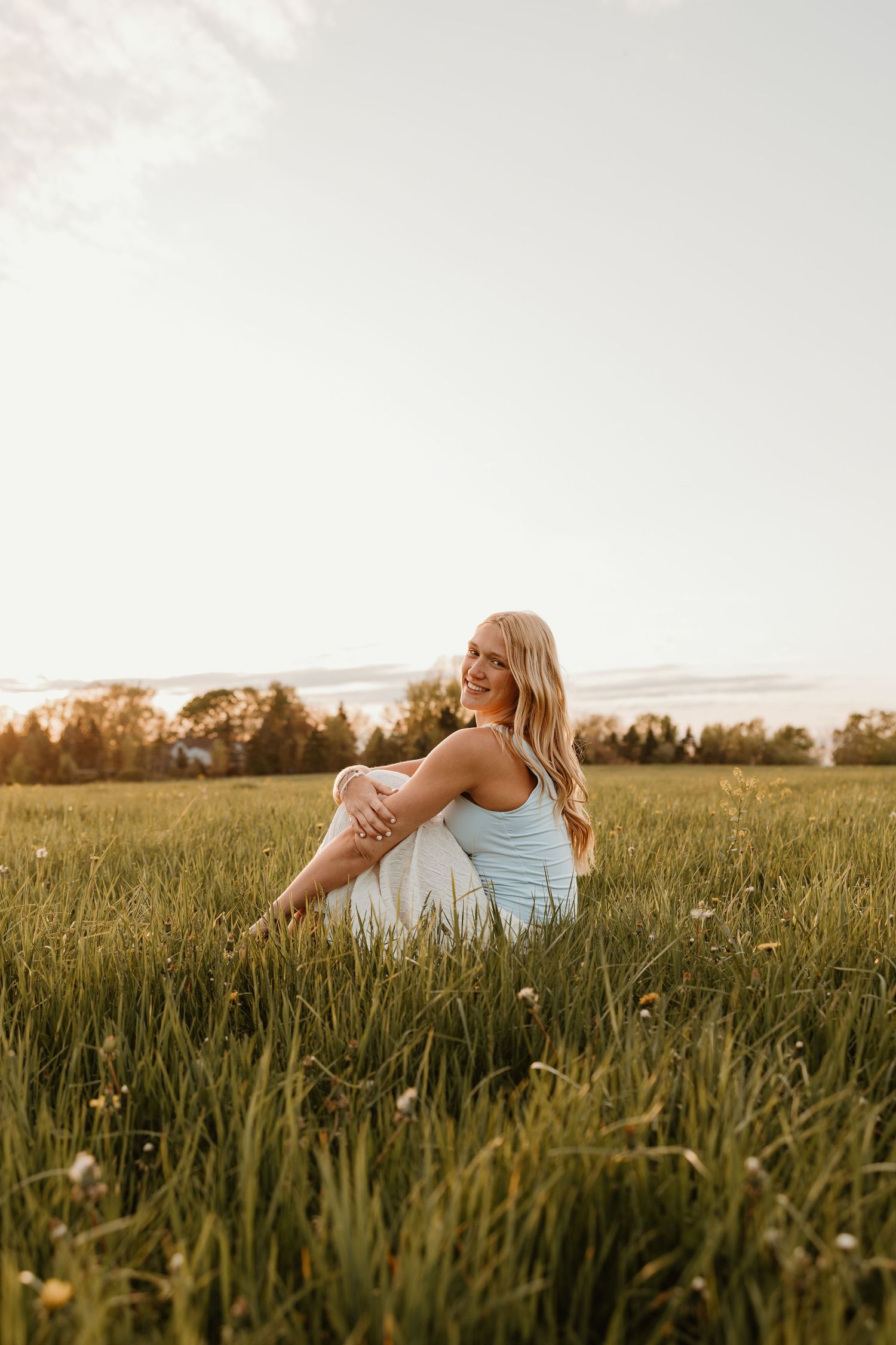 Smiling blonde woman sitting in a lush green meadow with wildflowers at golden hour sunset.